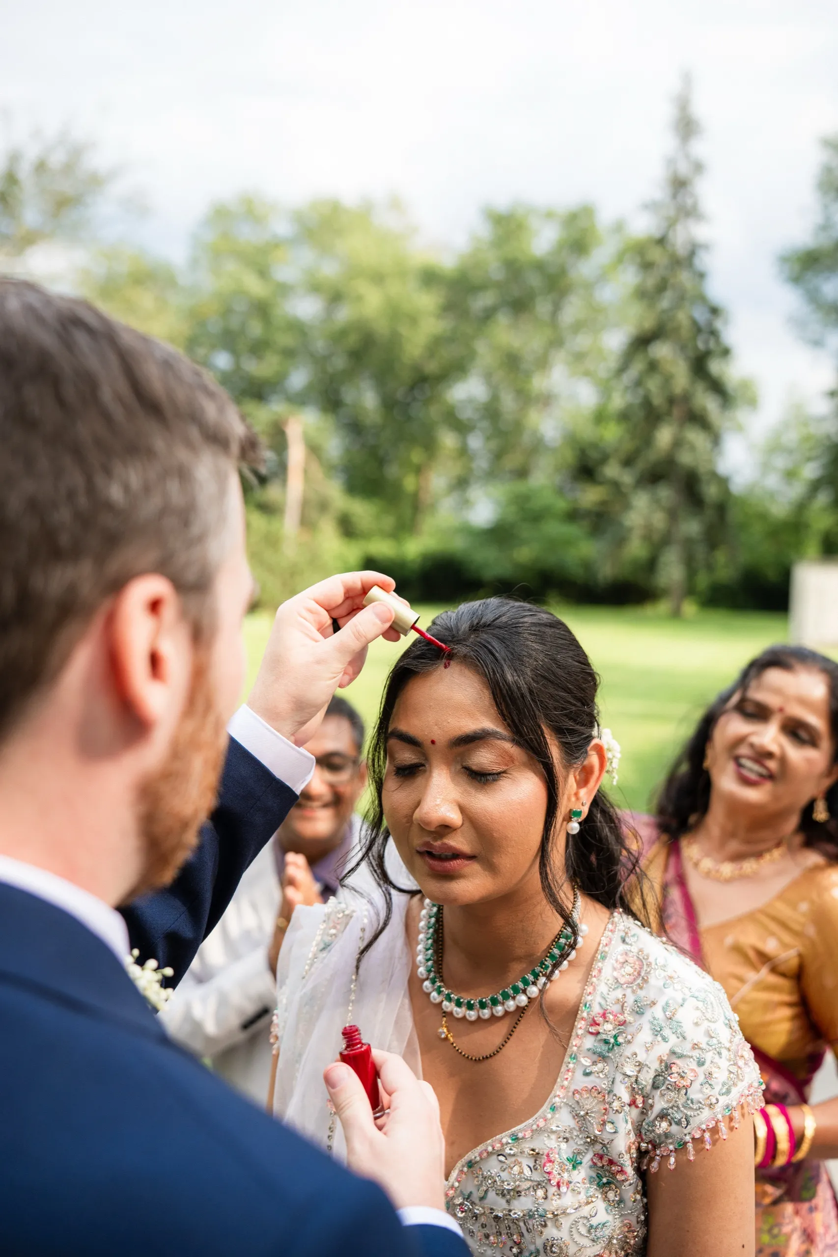 Derek marks Nidhi's forehead with a tilak as her parents smile through the ritual., by AD Photography