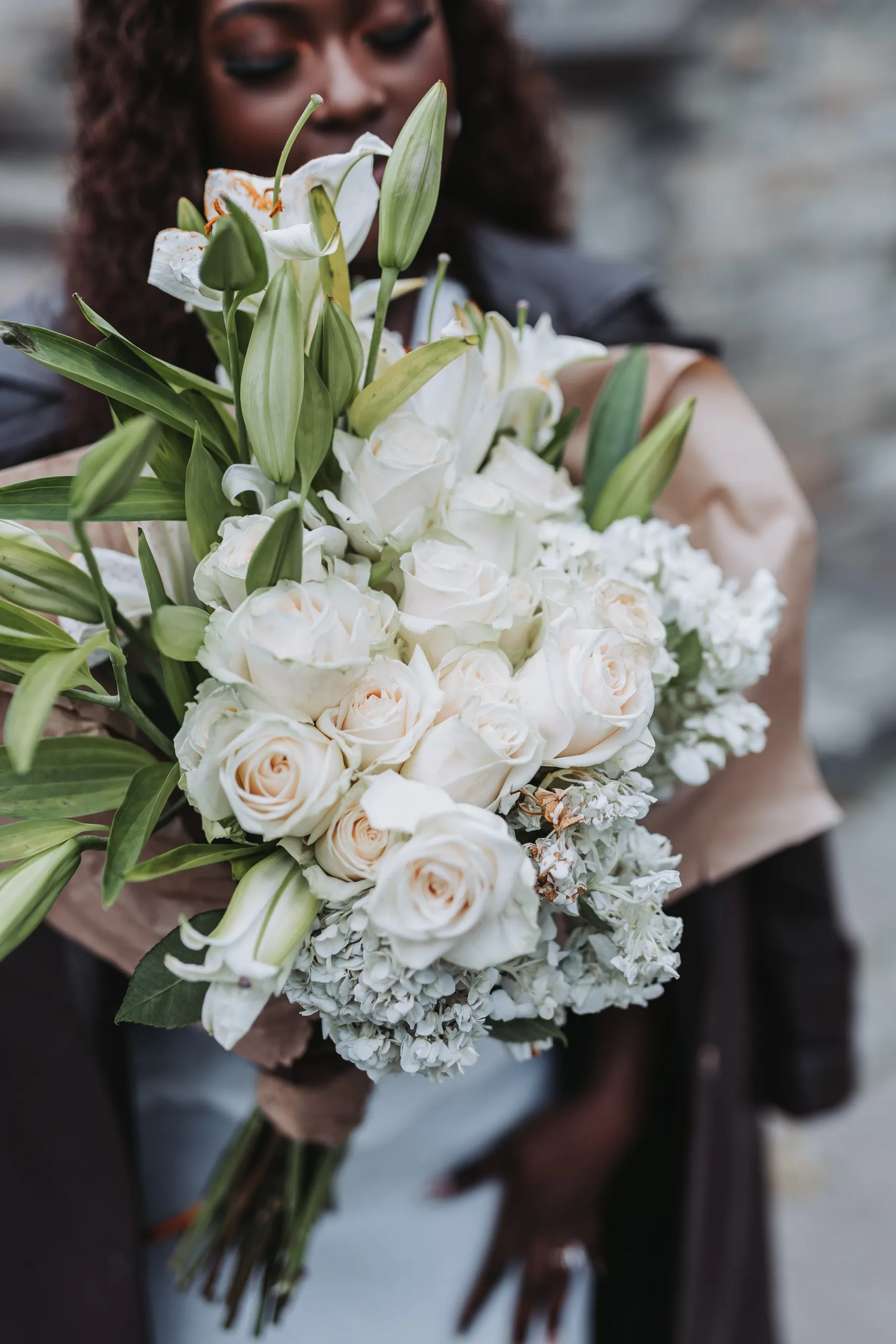 A bouquet of white roses and lilies held to the camera with Aimee's face soft behind it, by AD Photography