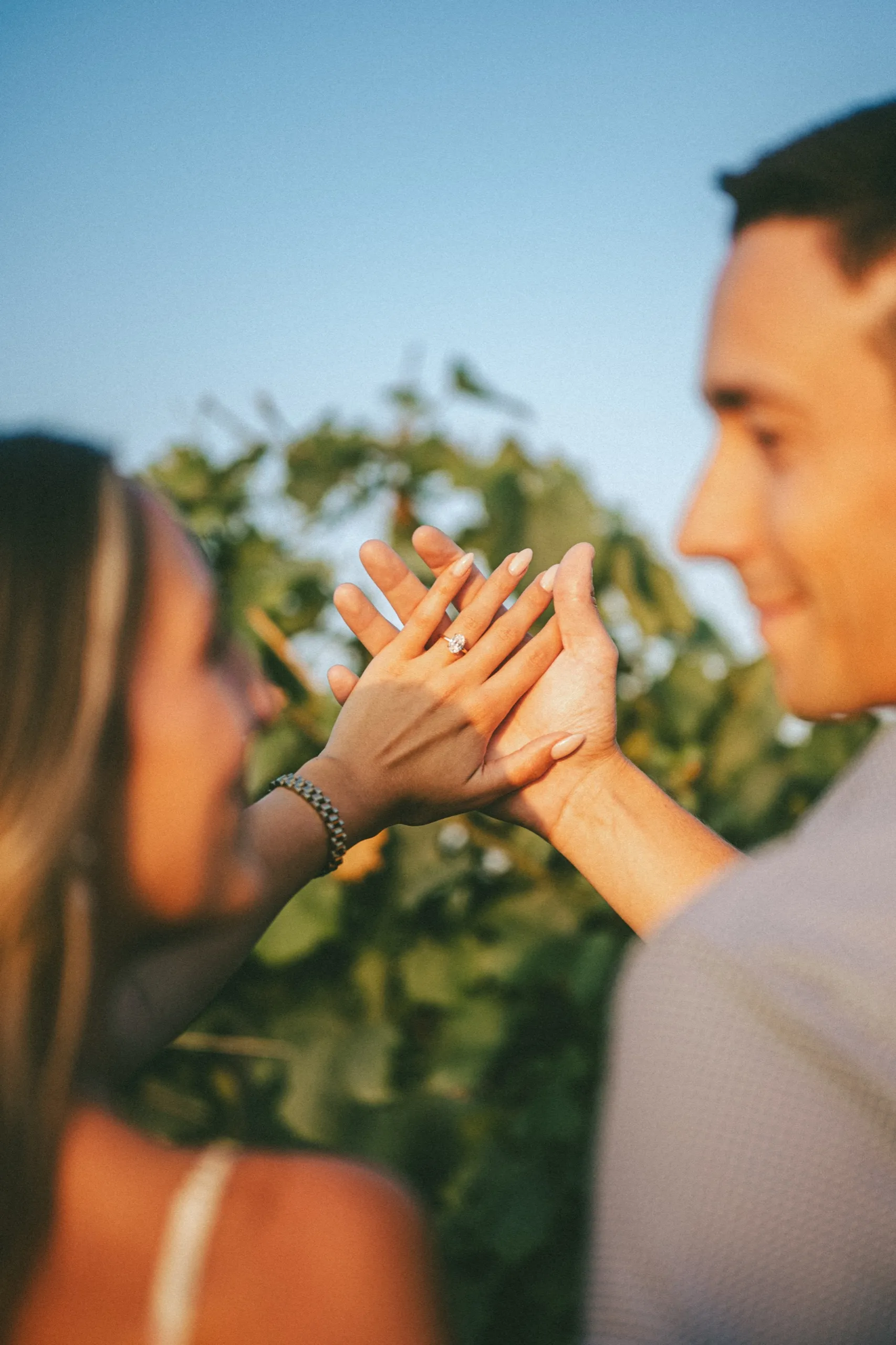 Ashley's engagement ring between their palms, held up against the sky over the vines, by AD Photography
