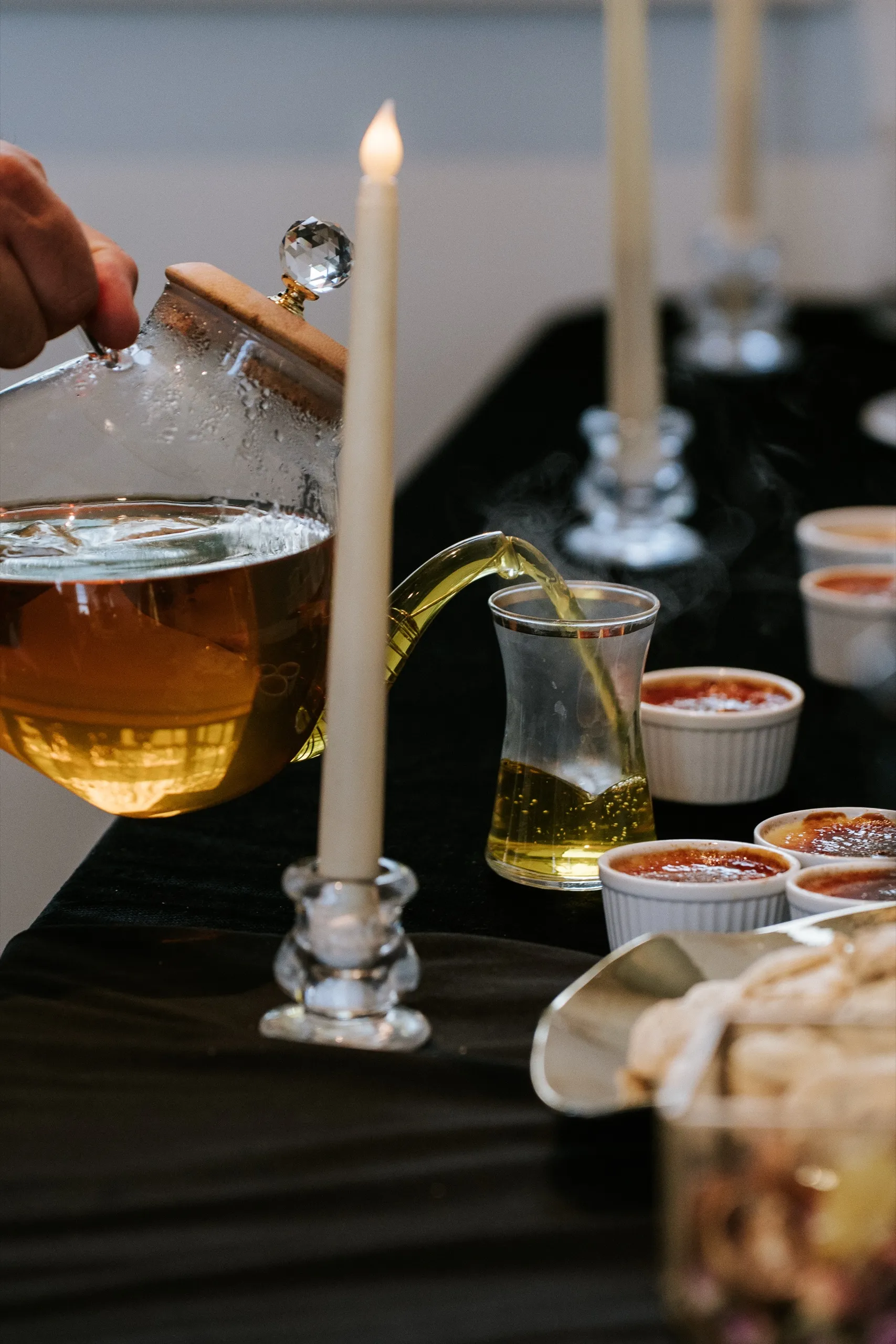 Tea poured from a glass pot into a small glass beside taper candles and creme brulee, by AD Photography