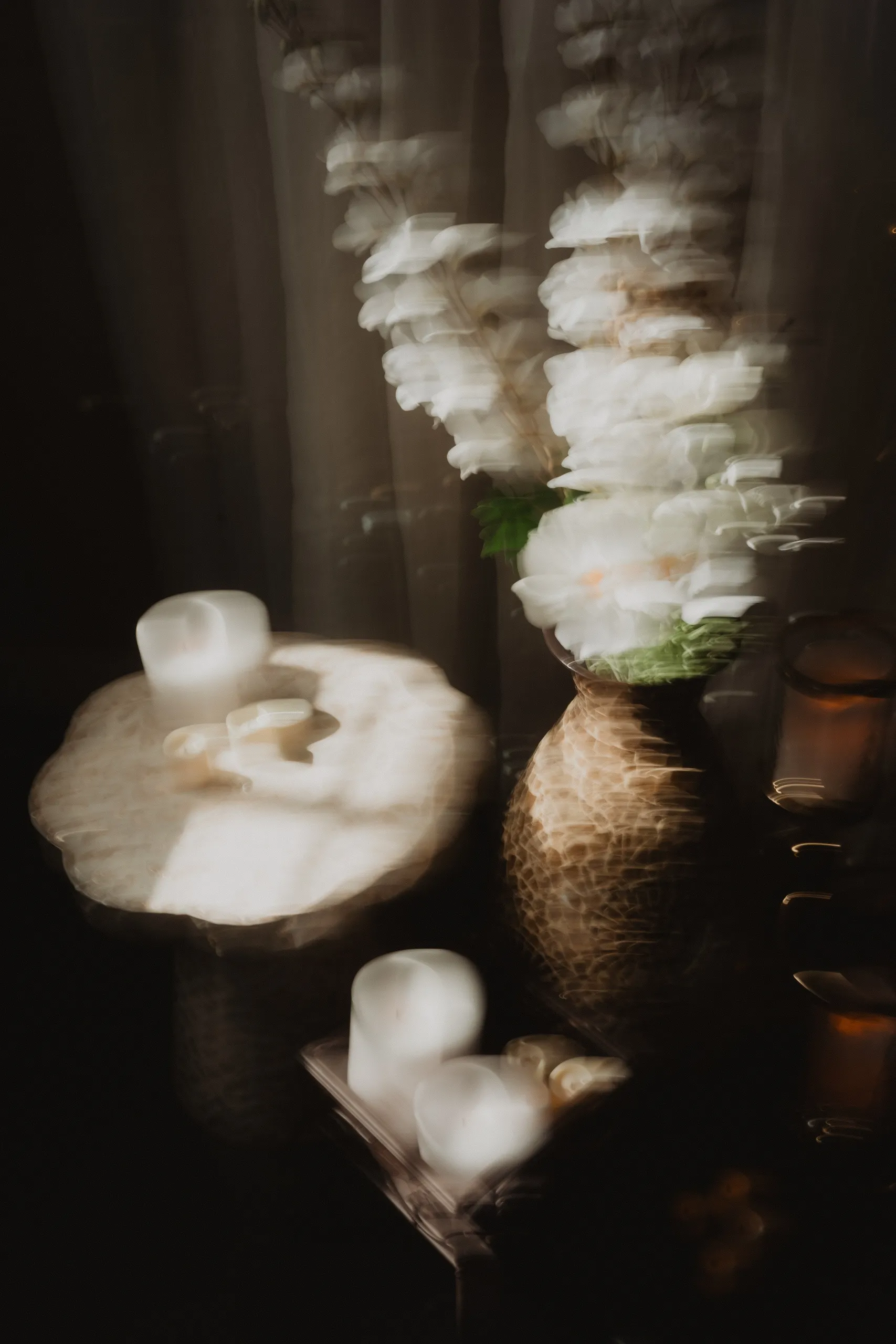 Long exposure blur of white blooms and candlelight glowing beside a sheer curtain, by AD Photography