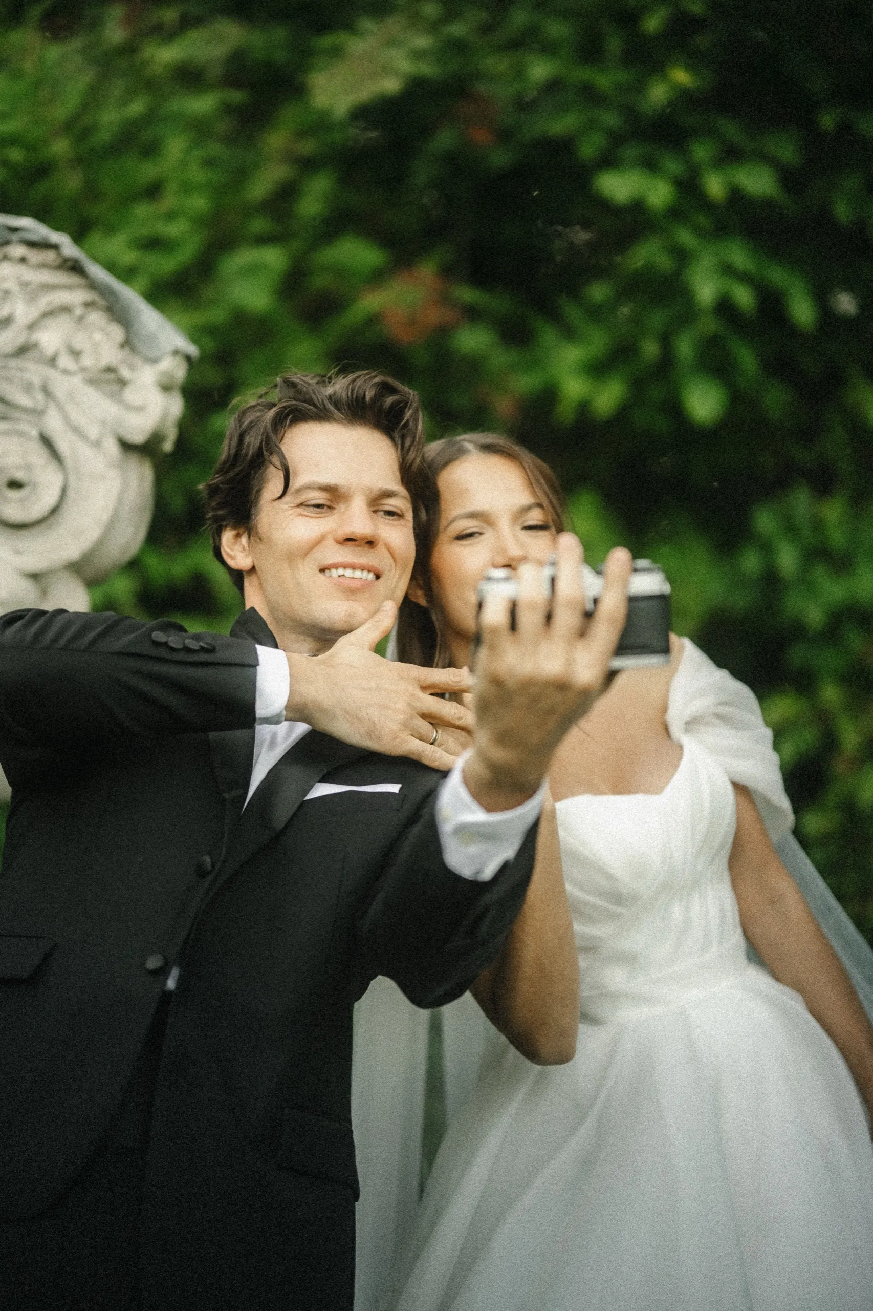 Groom holding up a vintage film camera for a selfie with the bride beside a carved stone urn, by AD Photography