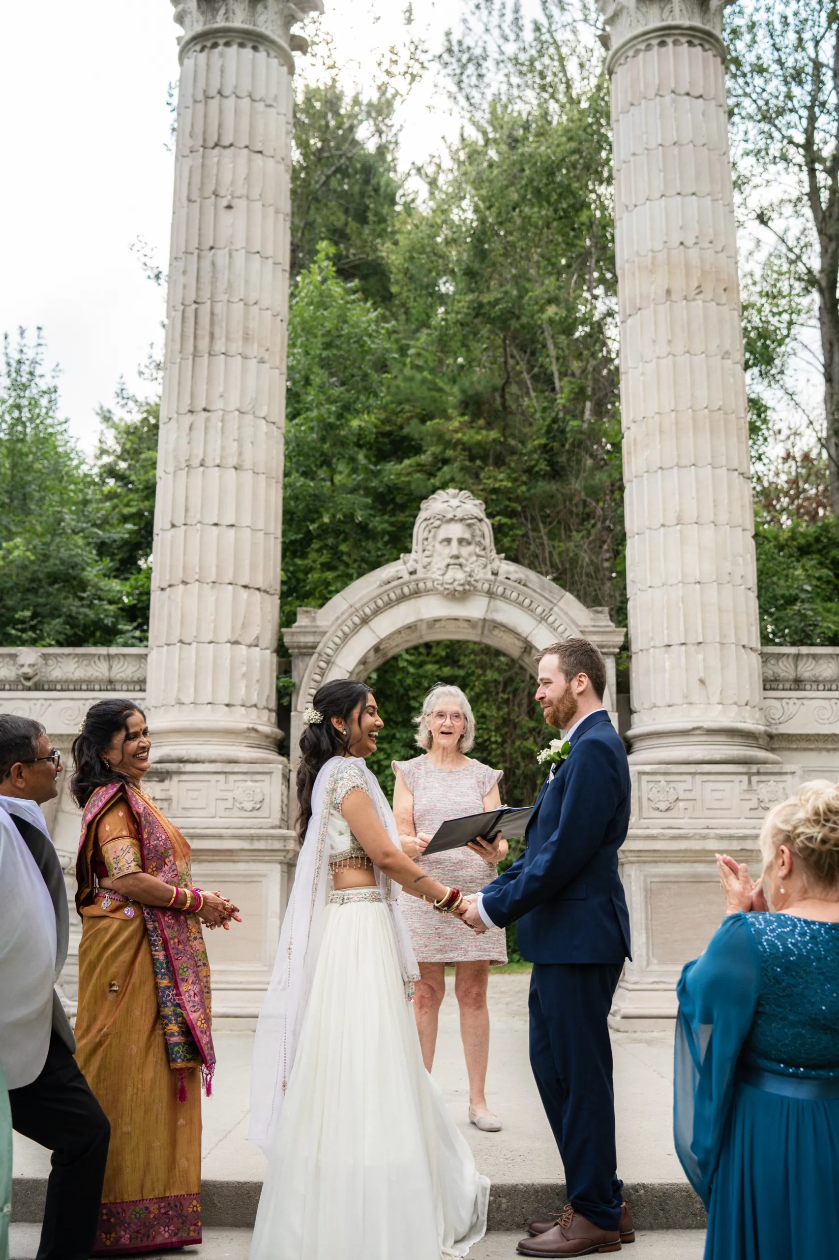 Nidhi and Derek hold hands laughing as the officiant reads on, family close beside them., by AD Photography