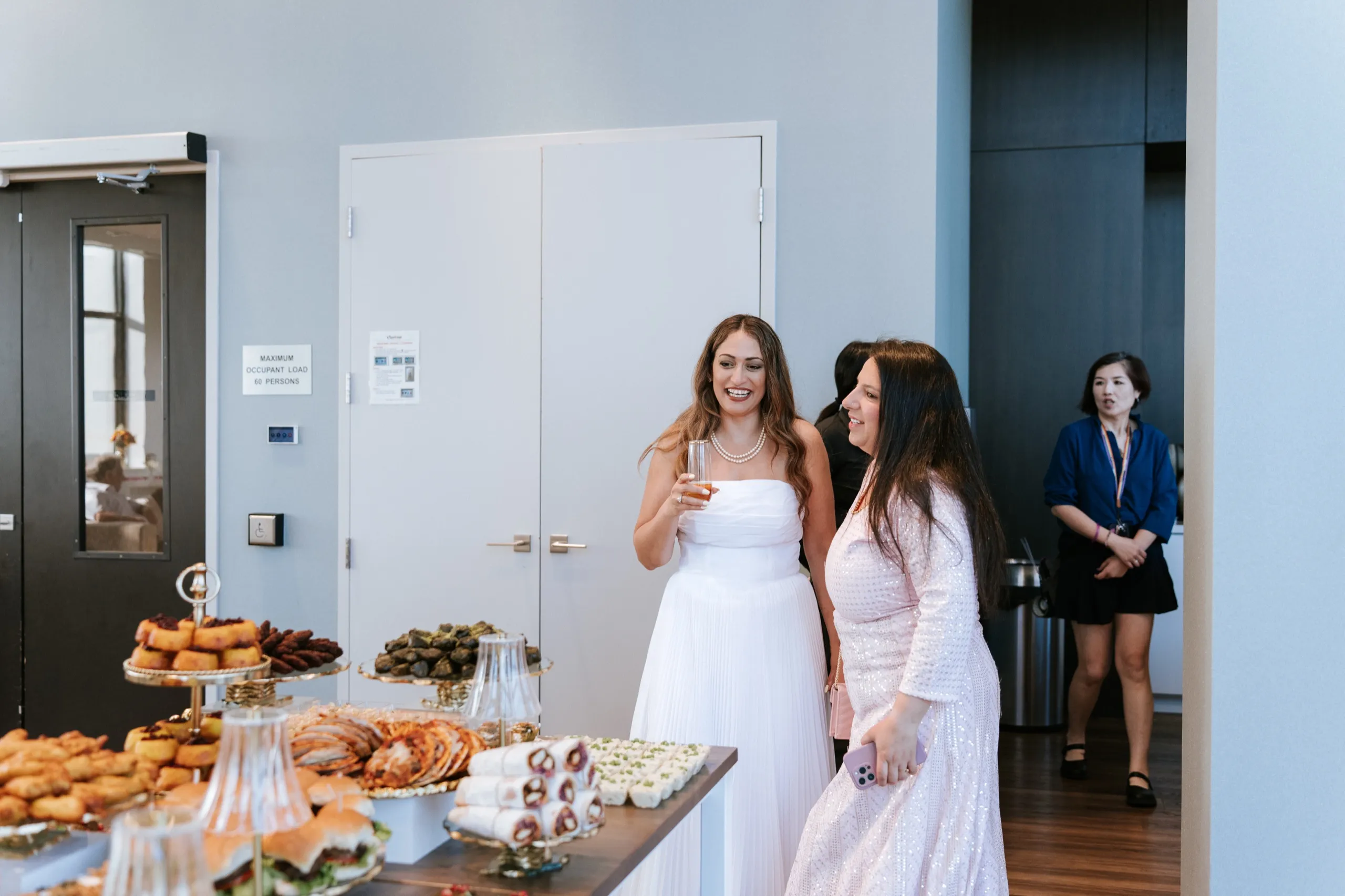 Two guests laugh together by the doors, seen past the sandwiches and pastries on the table, by AD Photography