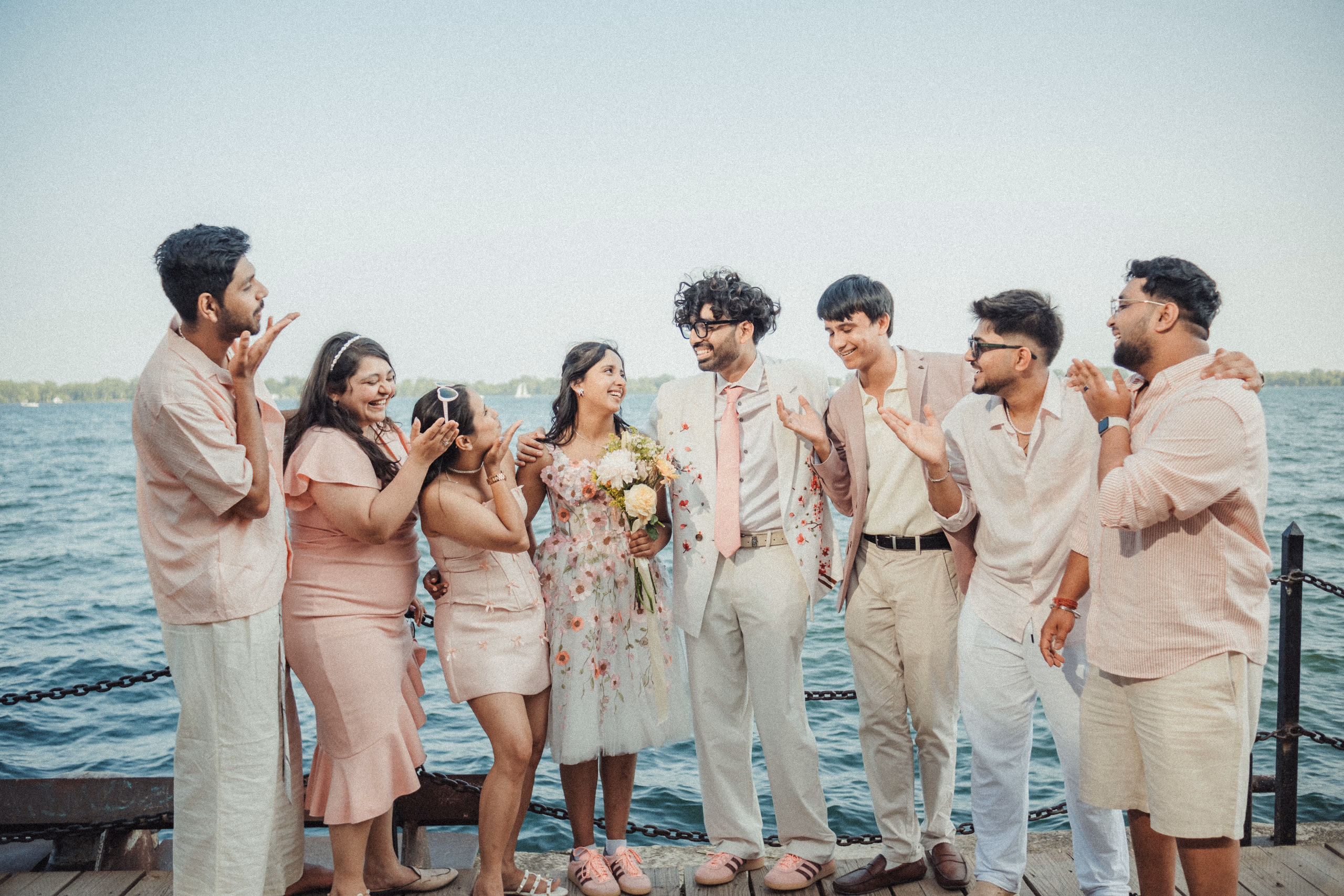 Harsh and Payal laughing with seven friends dressed in blush and cream on a Toronto Islands pier, the lake and a distant sailboat behind them, by AD Photography