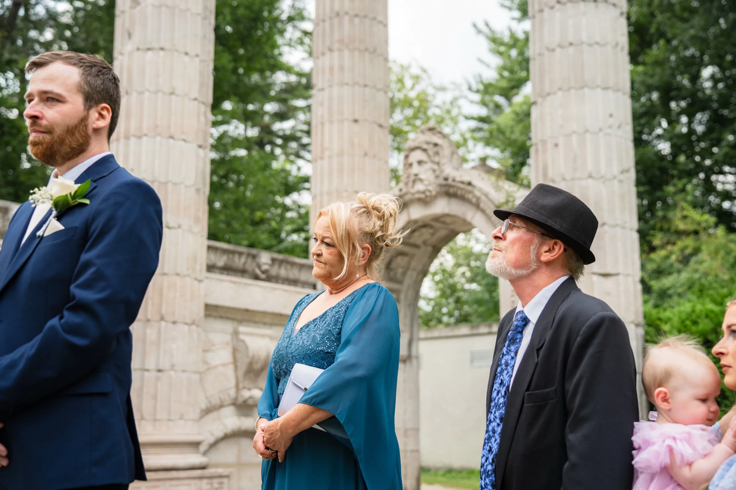 Derek's parents watch the vows, his father's gaze drifting up to the carved stone face., by AD Photography