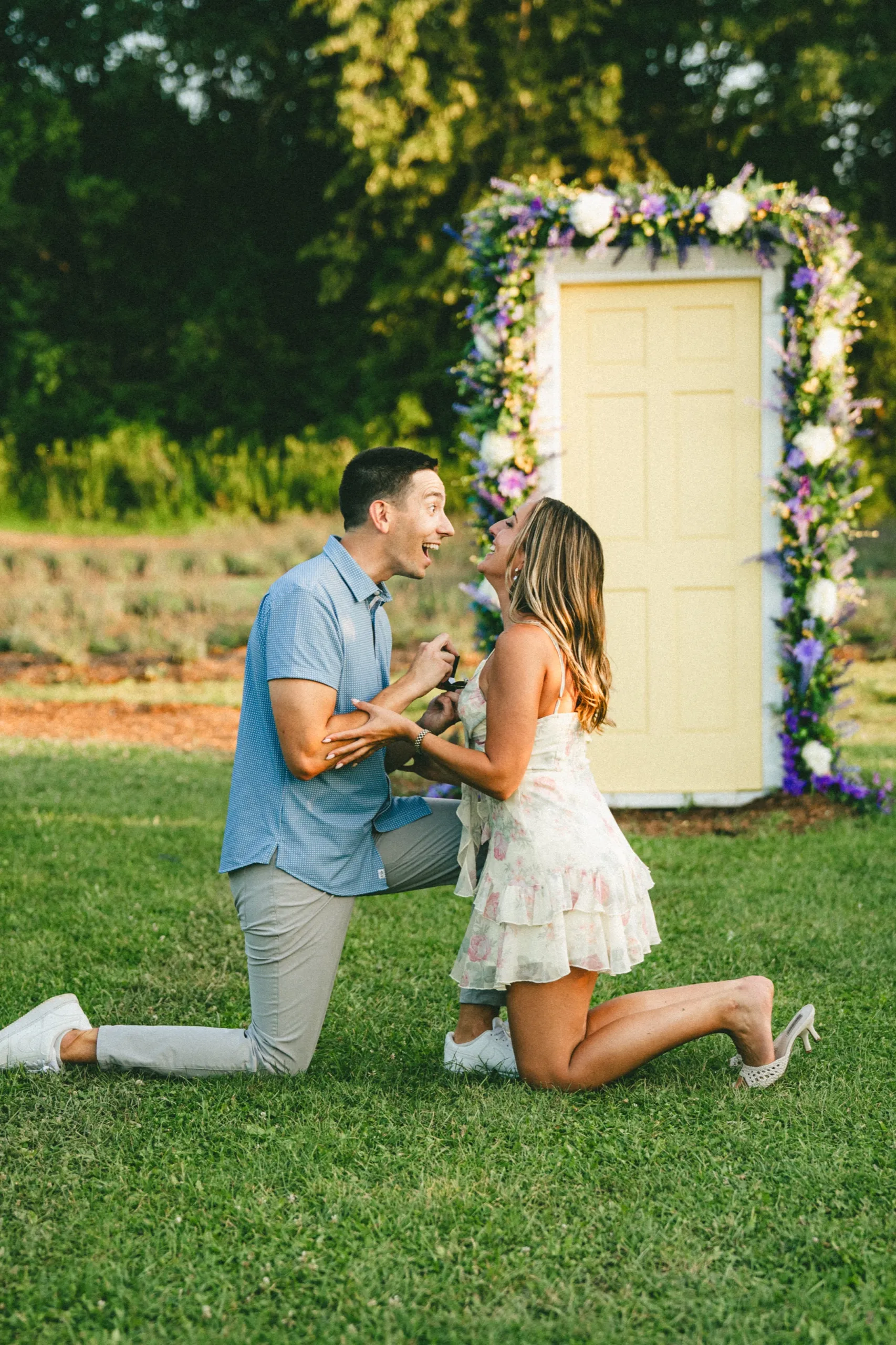 Both of them down on the grass, laughing, in front of the lavender and white floral arch, by AD Photography