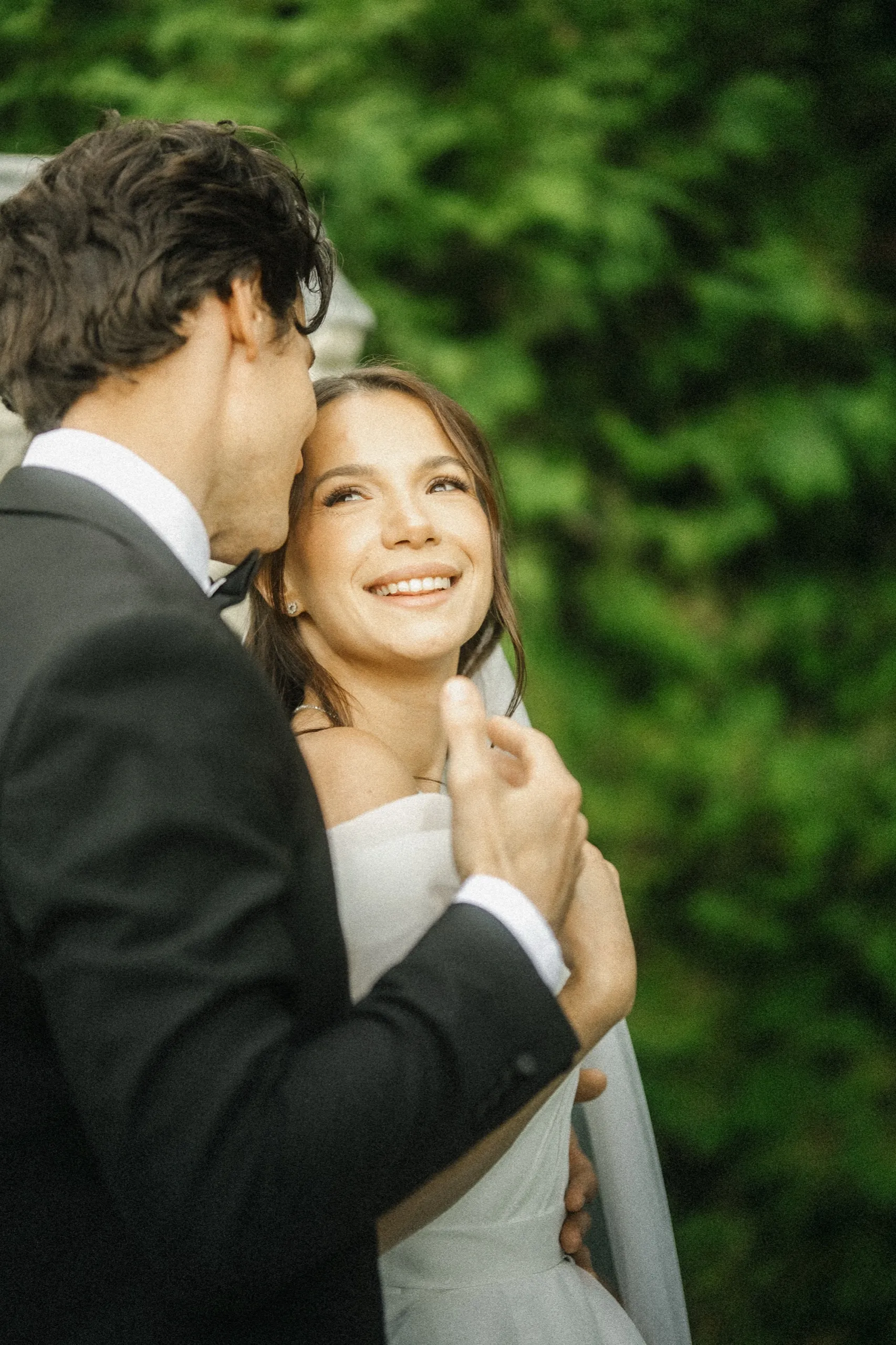 Bride laughing as the groom buries his face in her hair against a green hedge, by AD Photography
