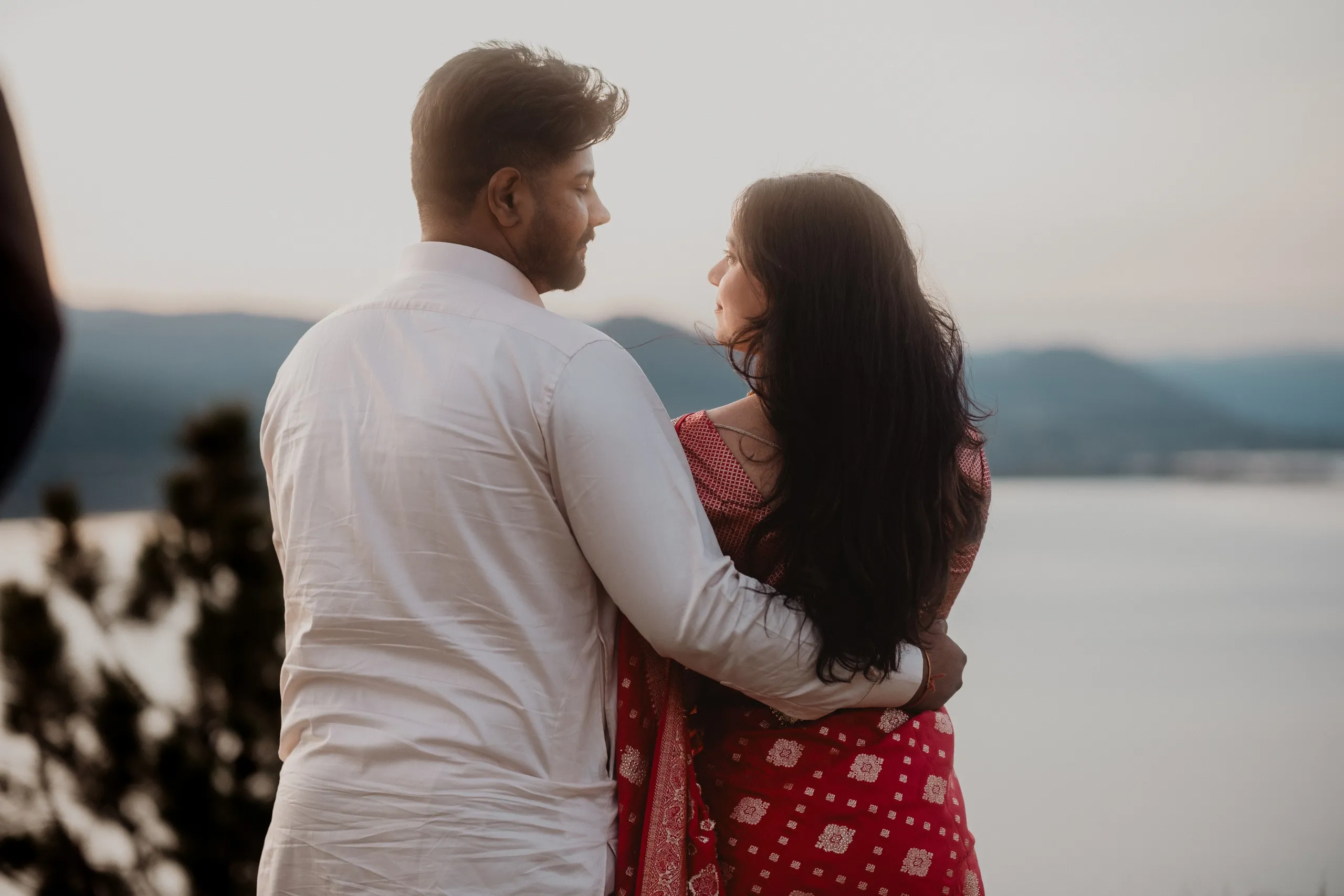 Golden hour on a lakeside bluff, Mehak in a red saree standing close to Kamal, by AD Photography