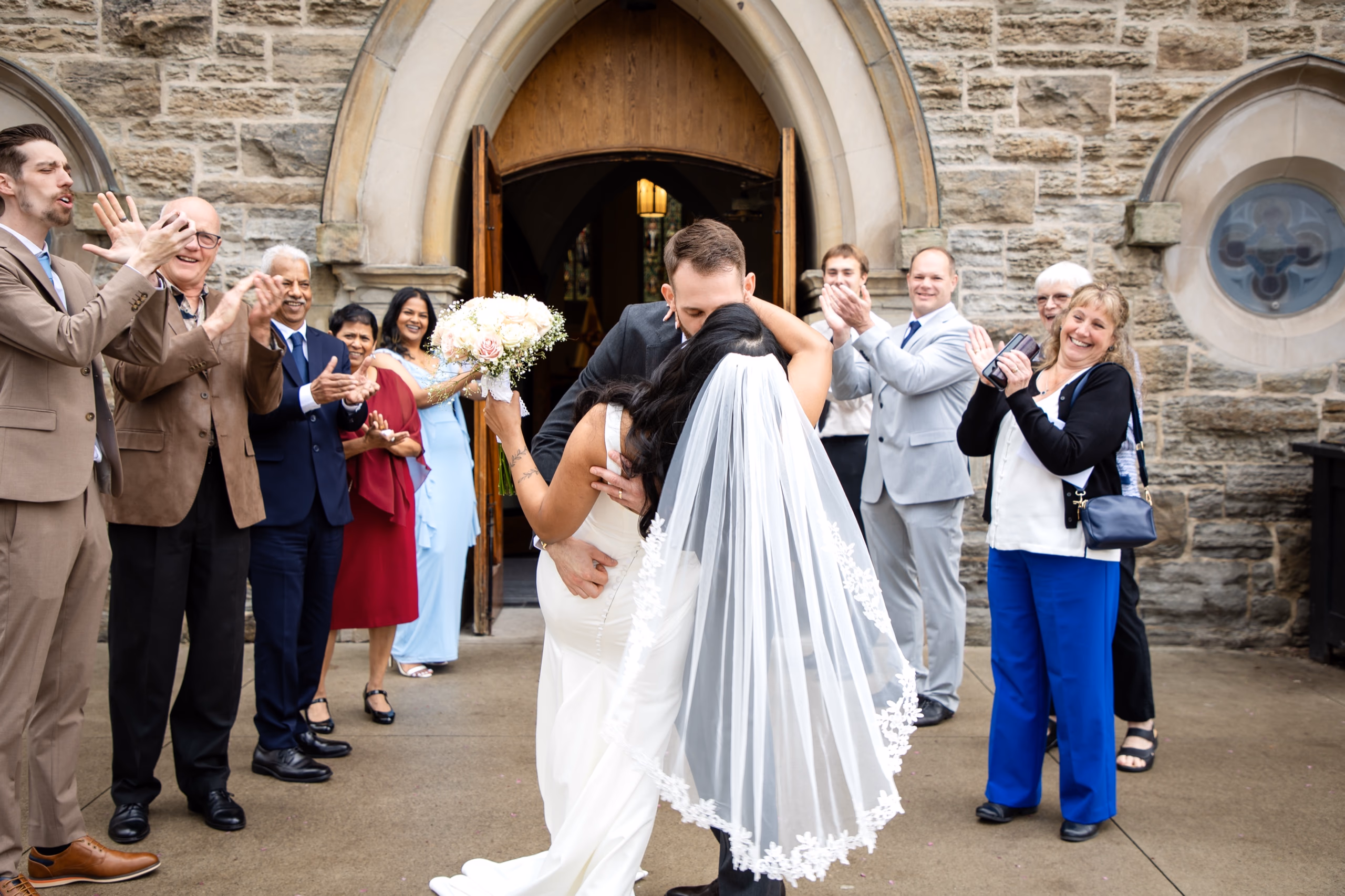 Roxanne and Justin embrace on the church steps of St. Patrick's Catholic Parish in Hamilton while family clap and cheer around them, featured by AD Photography