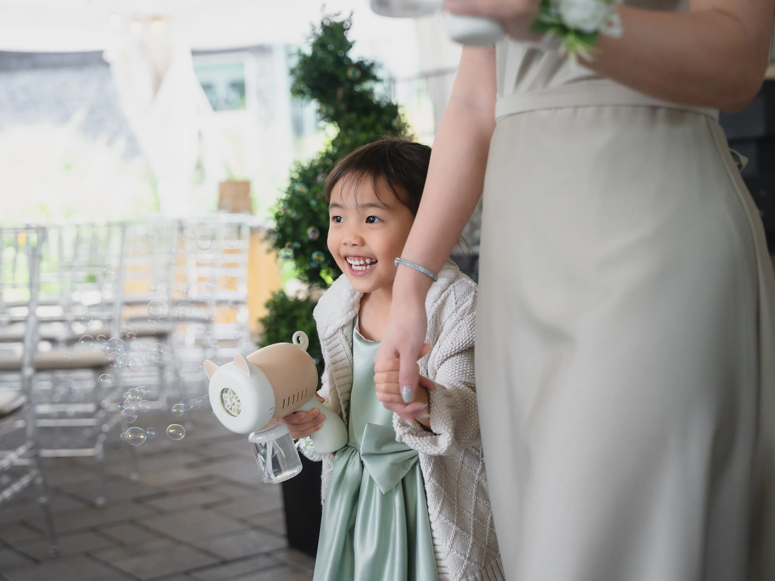 A flower girl in sage green firing a bubble gun before the Guild Park ceremony, by AD Photography