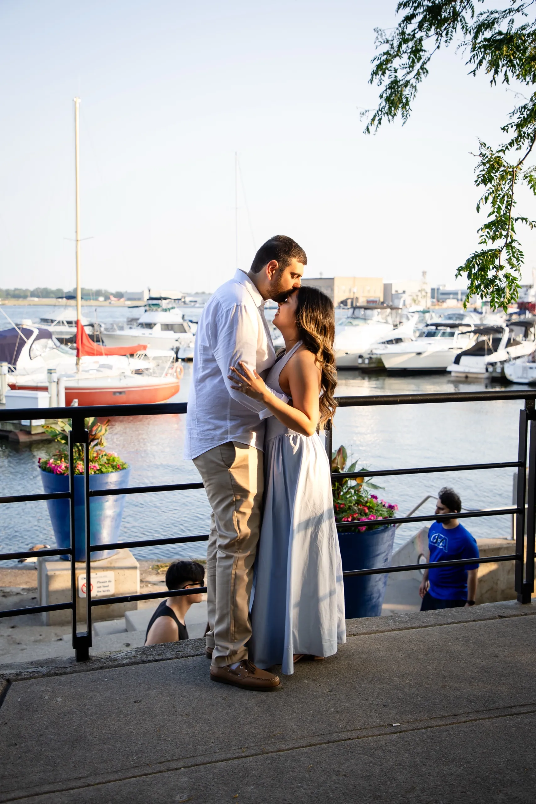 The engaged couple kissing at the marina railing beside the Toronto Music Garden, sailboats behind them, by AD Photography