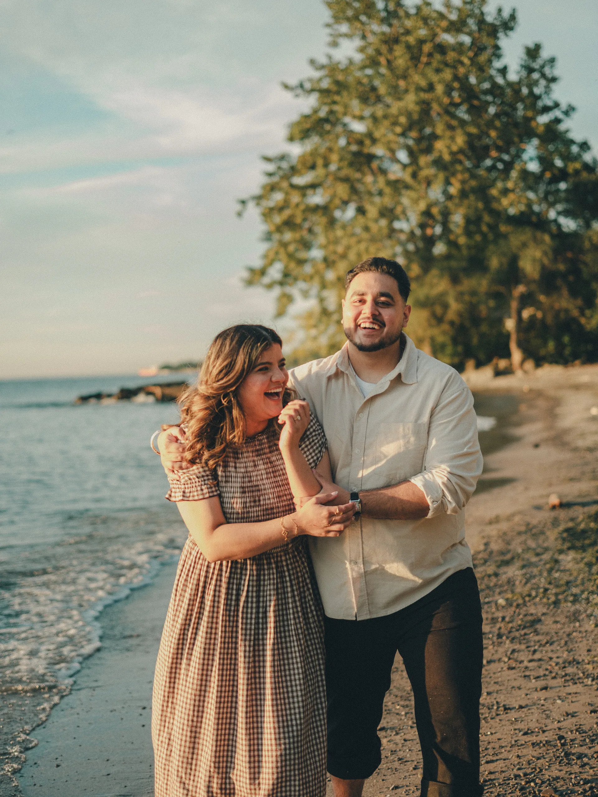 Parneet wrapped in Maninder's arm, both laughing under the big shoreline tree on a Lake Ontario beach, by AD Photography