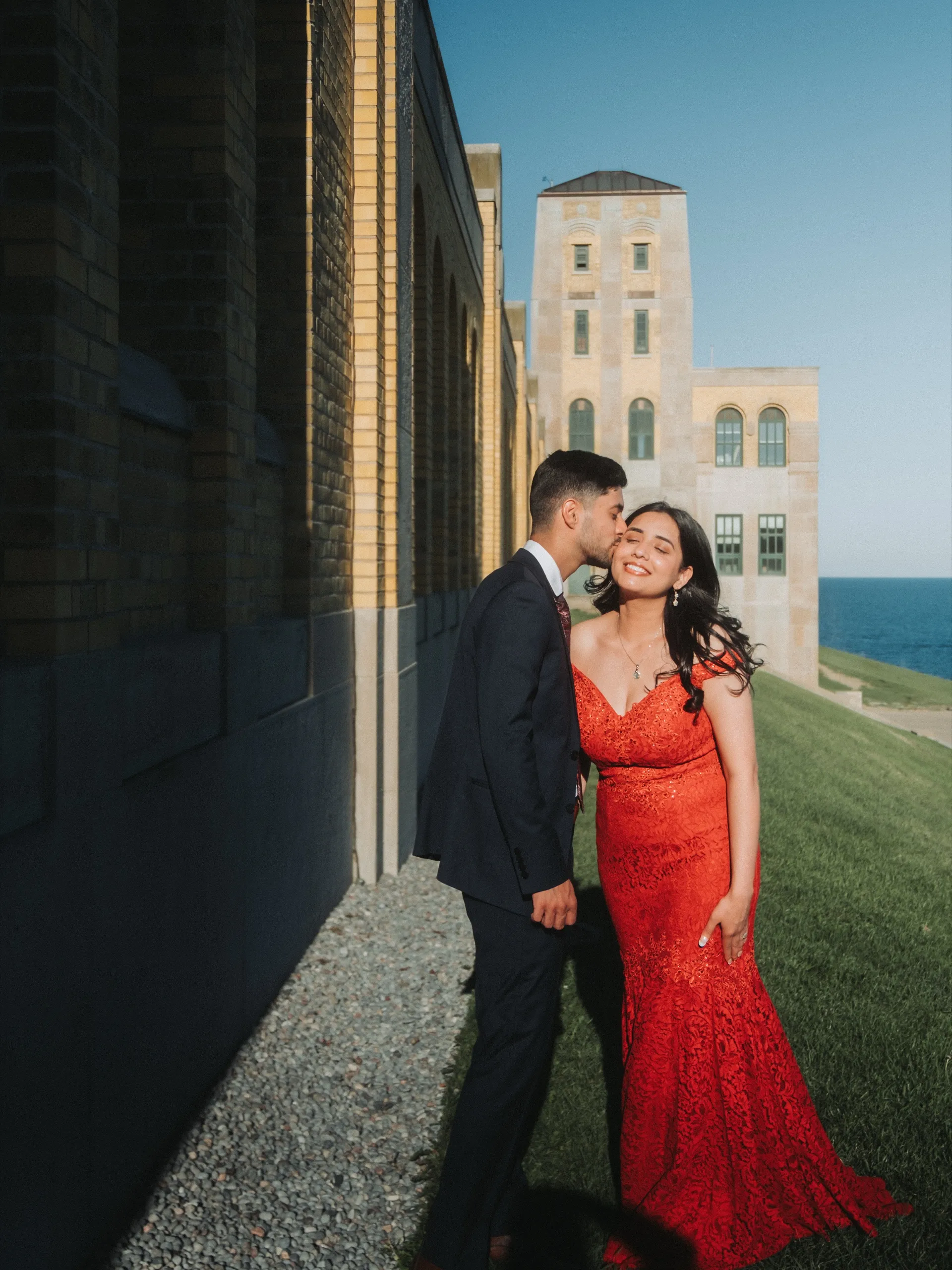 He kisses her cheek beneath the art deco tower with Lake Ontario beyond, by AD Photography