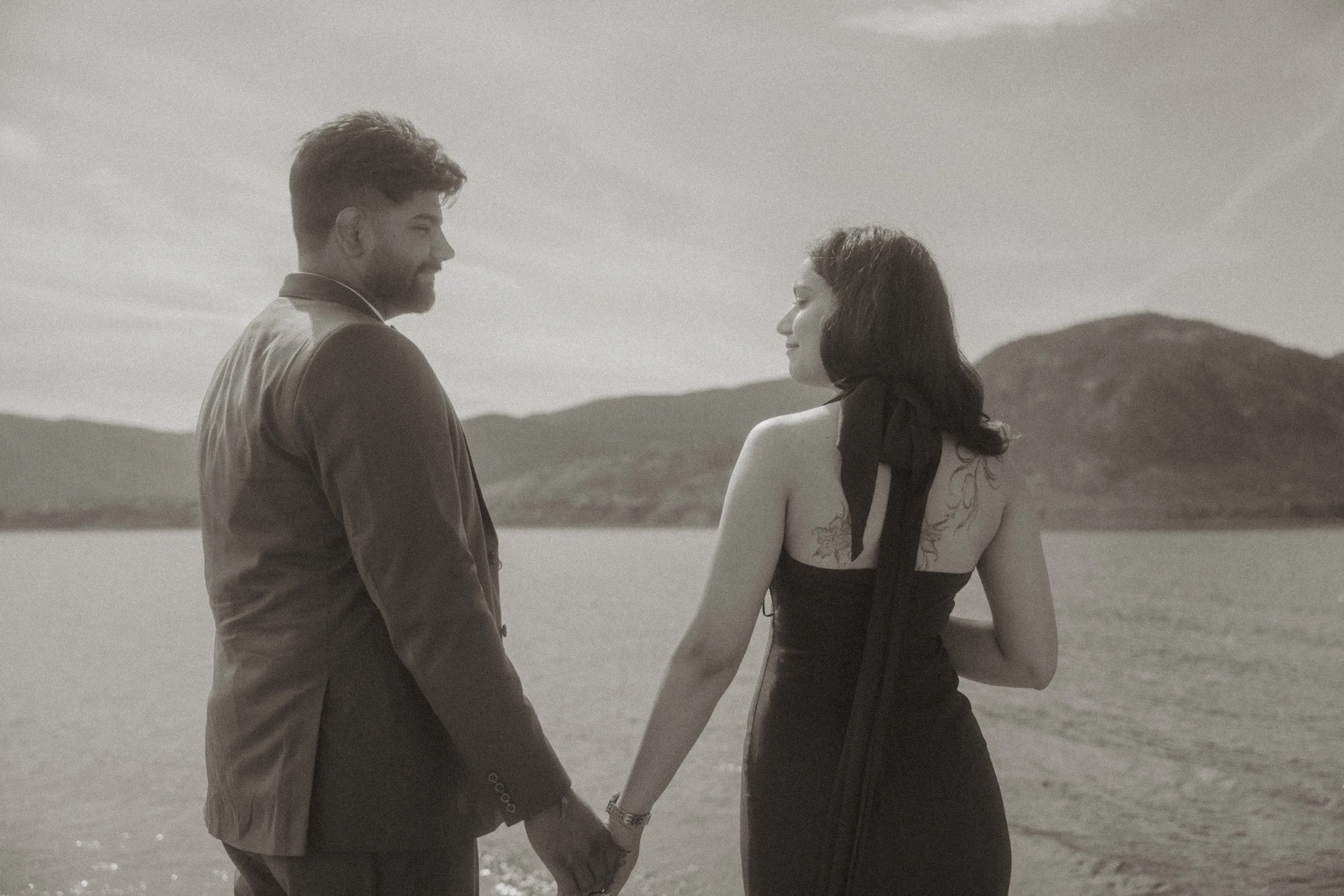 Black and white frame of the couple pausing hand in hand along the lakeshore, by AD Photography