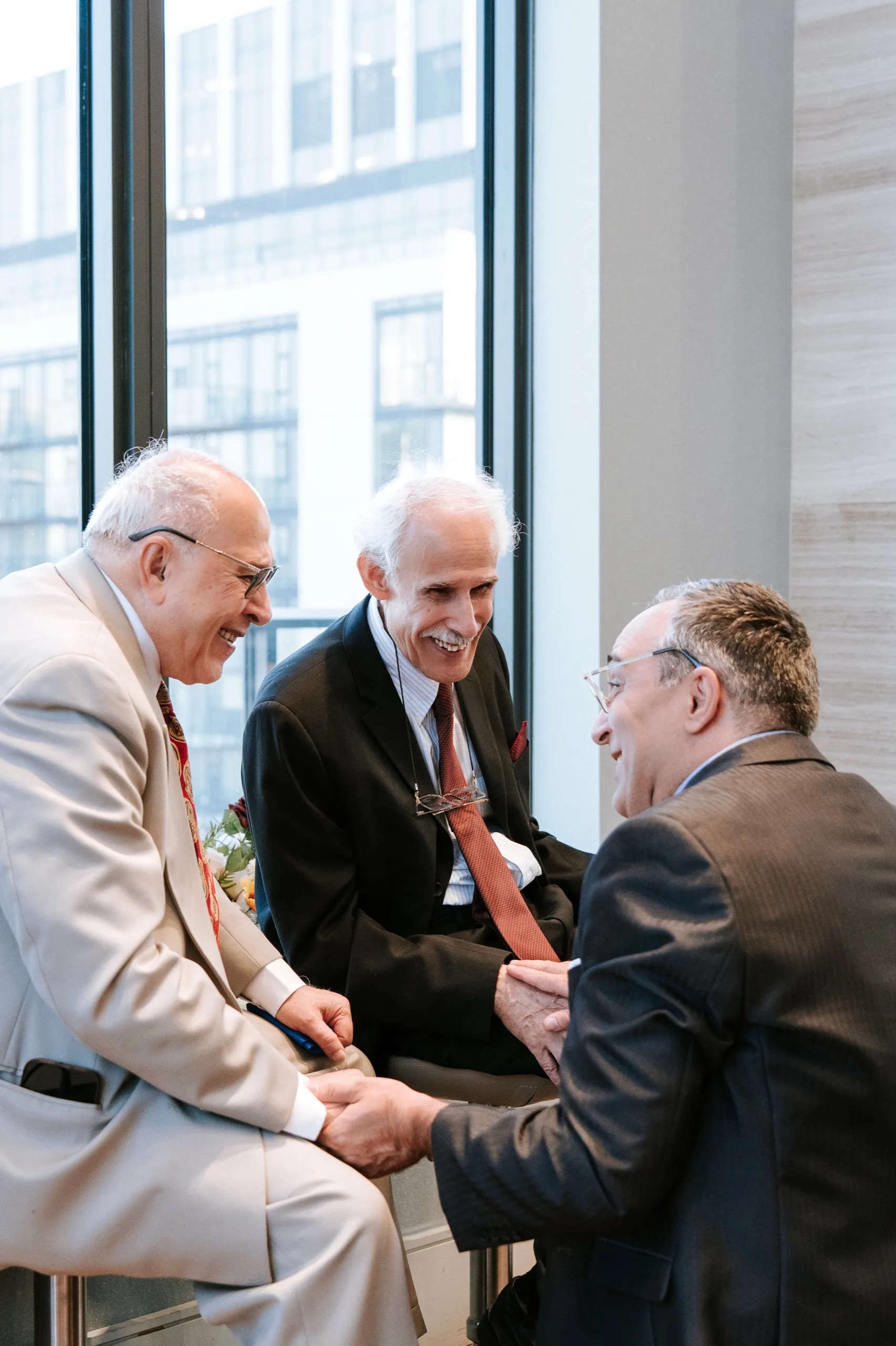 Three older guests shake hands and laugh together by the window, by AD Photography