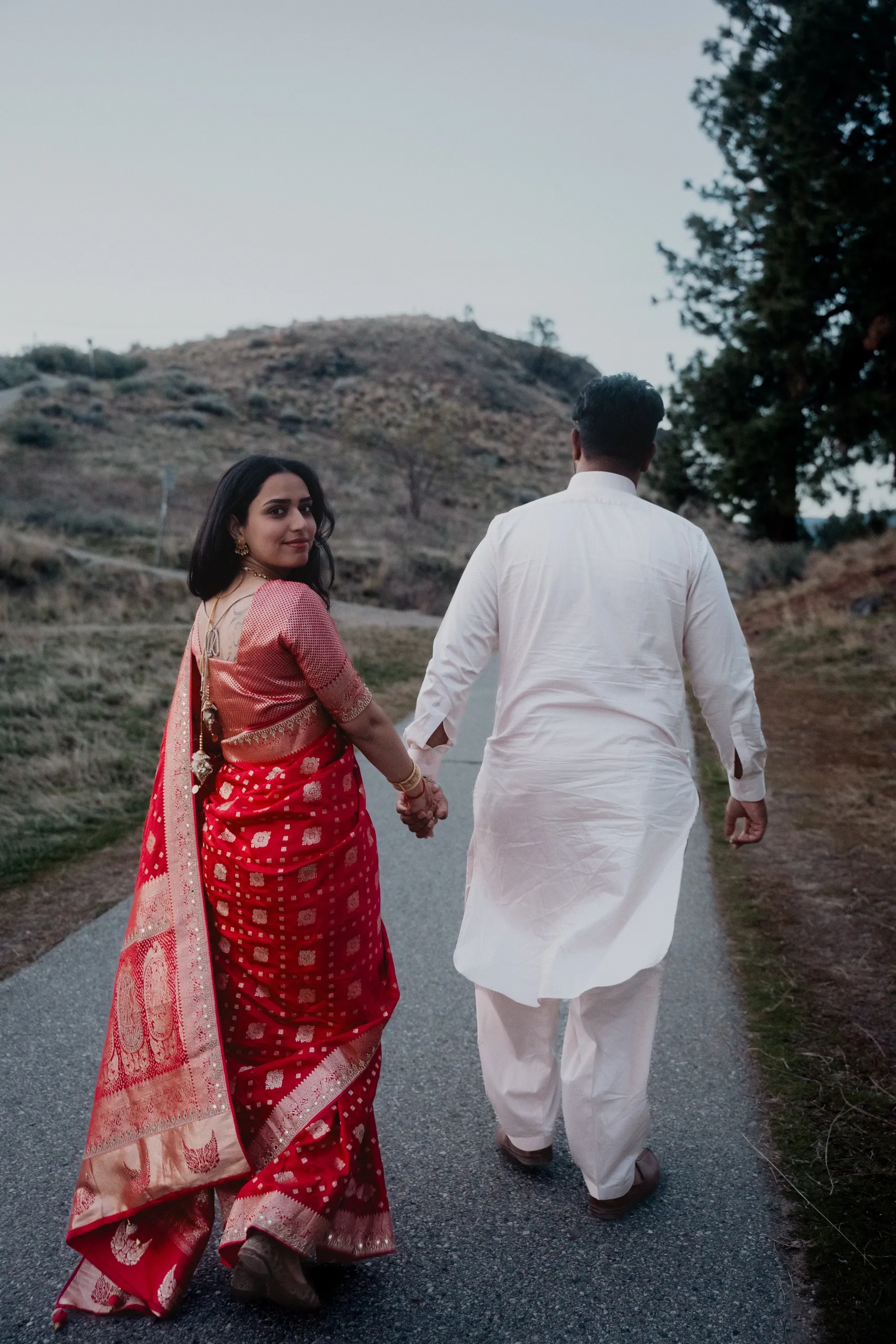 Mehak glancing back in her red saree while Kamal leads her up the farm road, by AD Photography