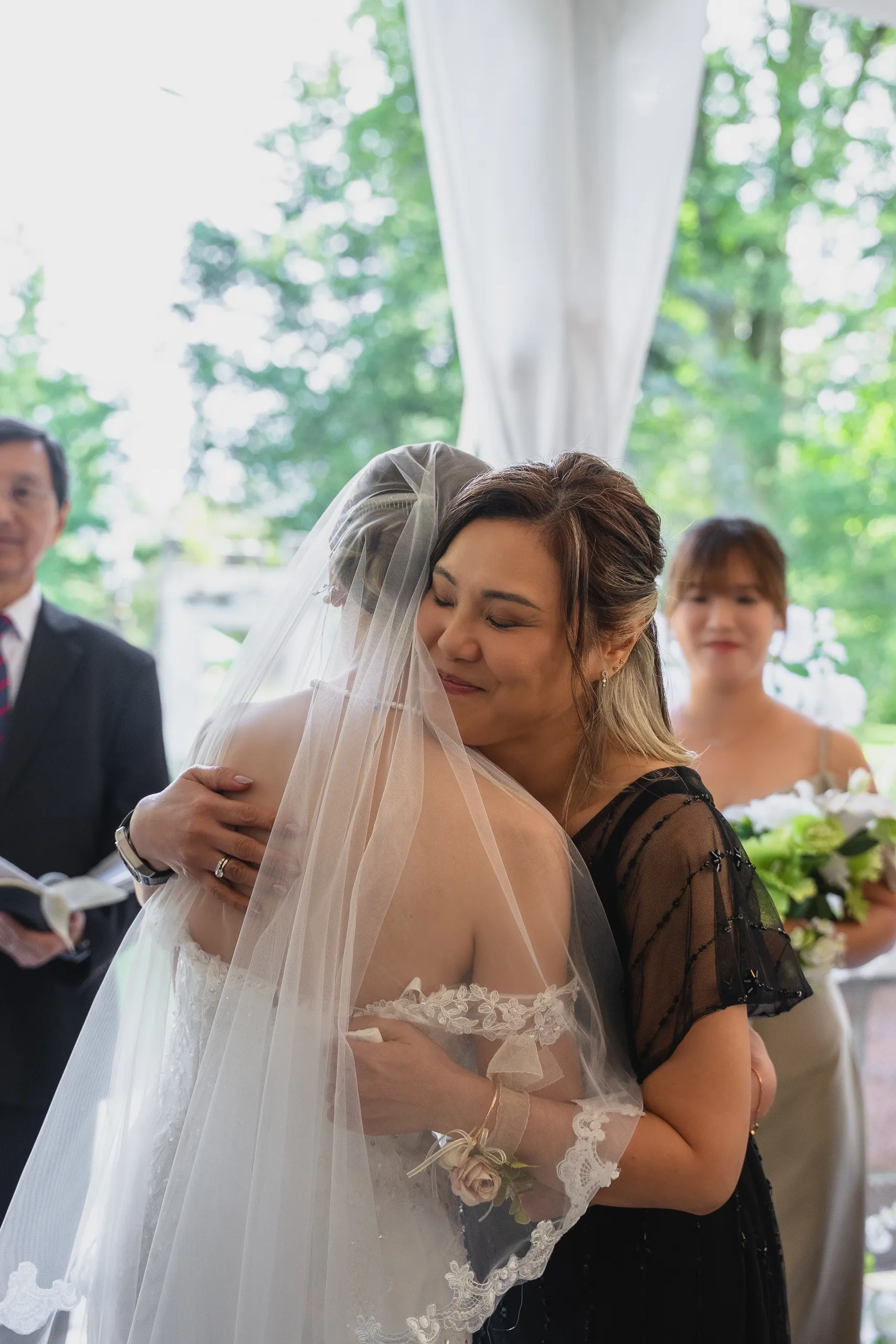 A guest in black lace holding veiled Josie in a long embrace at the ceremony, by AD Photography