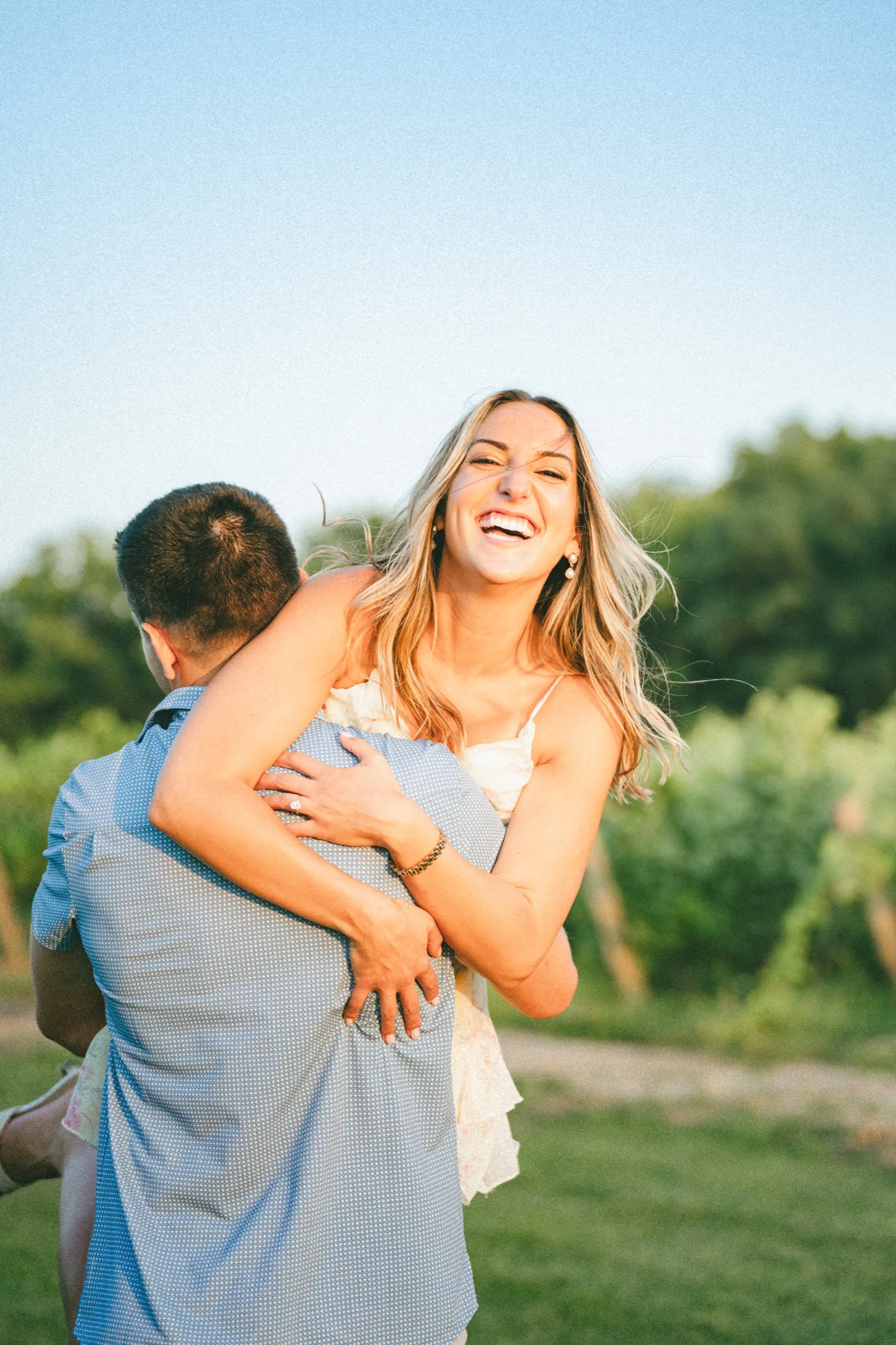Ashley laughing with her eyes shut as Donald holds her, vineyard and open sky behind, by AD Photography