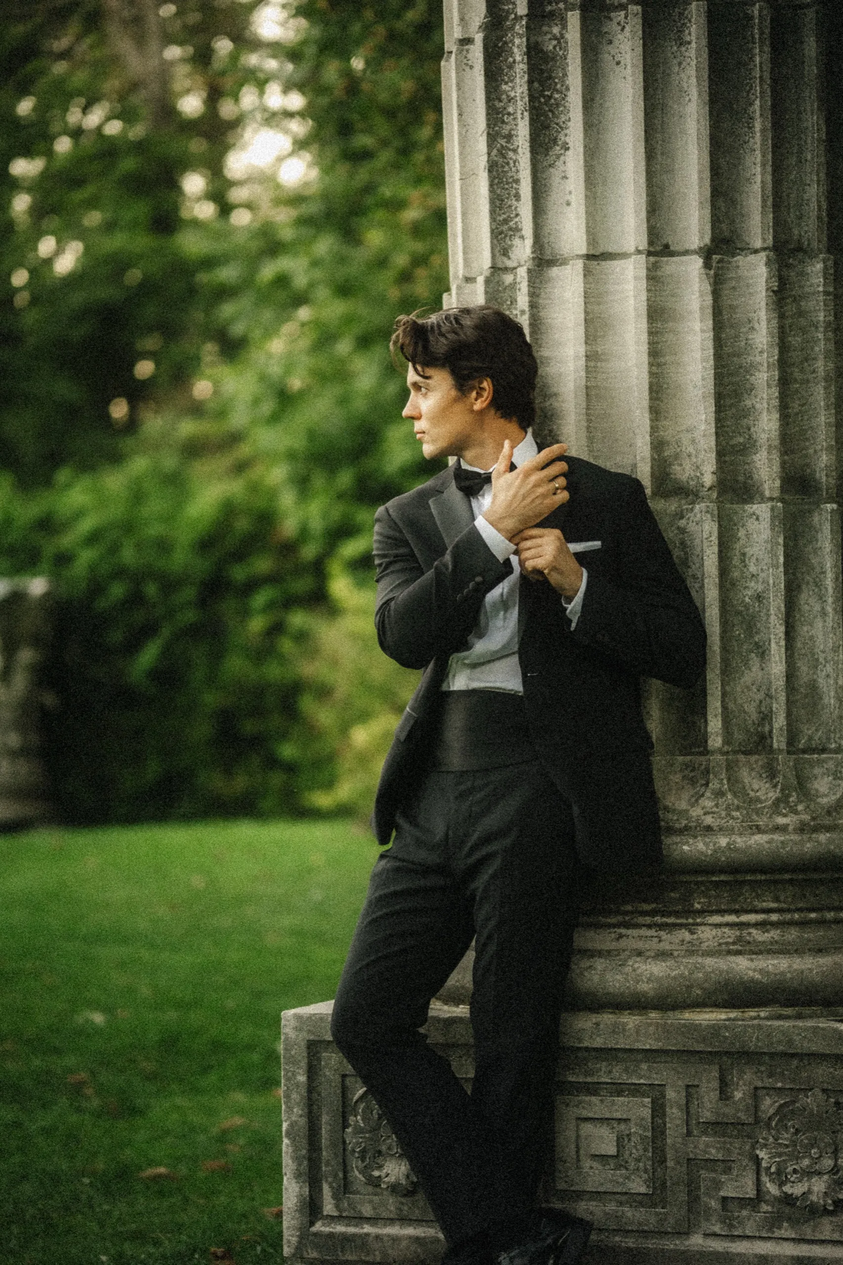 Groom adjusting his cufflink in profile, leaning on a stone column base, by AD Photography