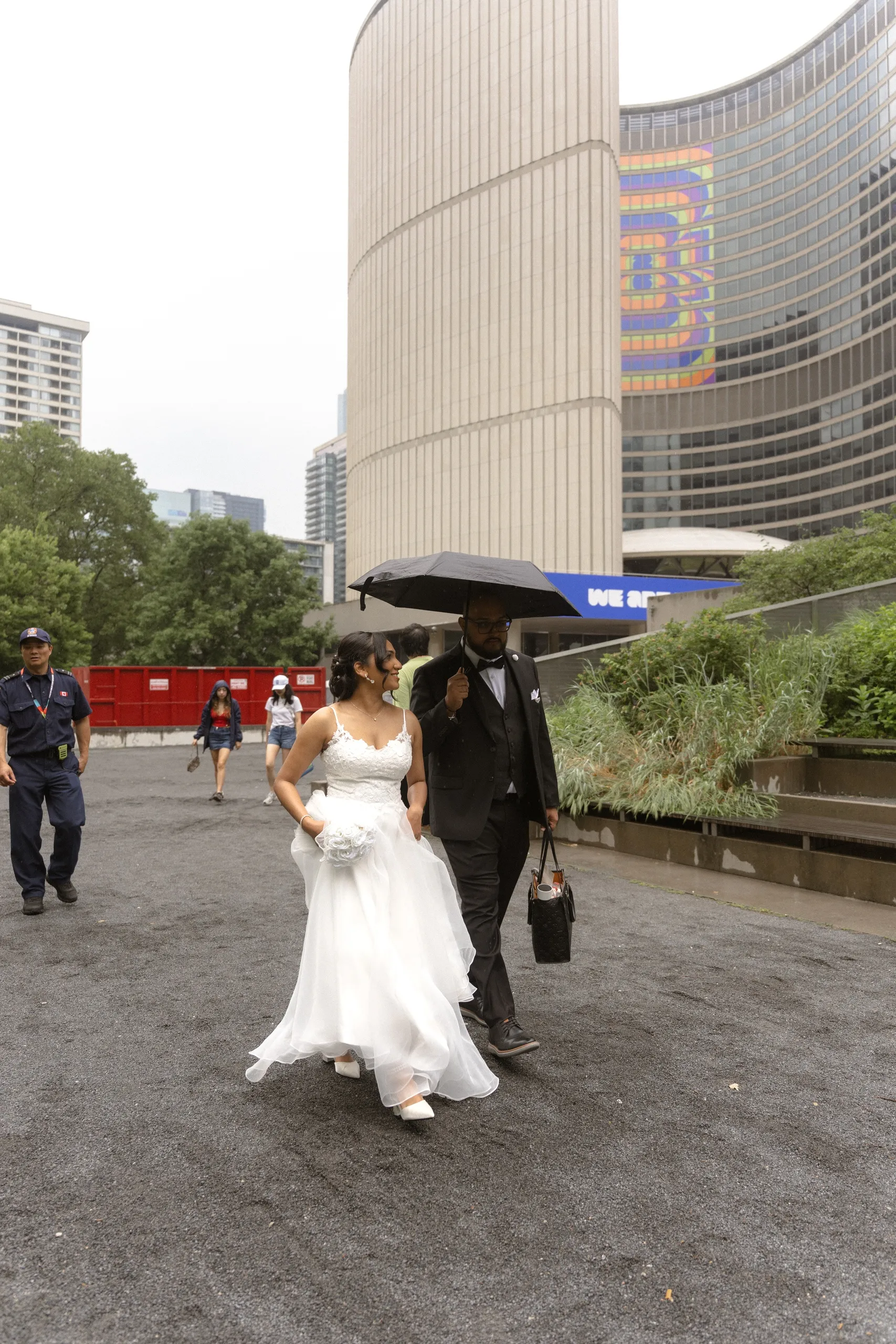 Bride and groom cross Nathan Phillips Square under an umbrella in the rain, by AD Photography