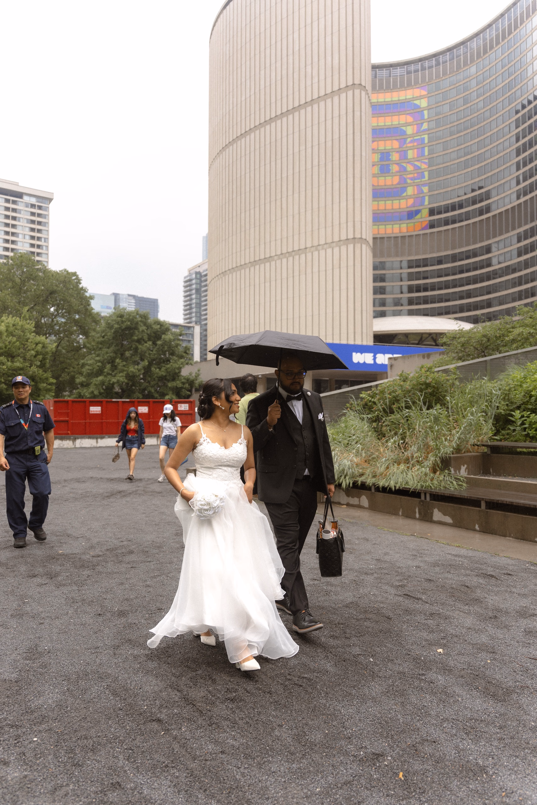 Bride and groom cross Nathan Phillips Square under an umbrella in the rain