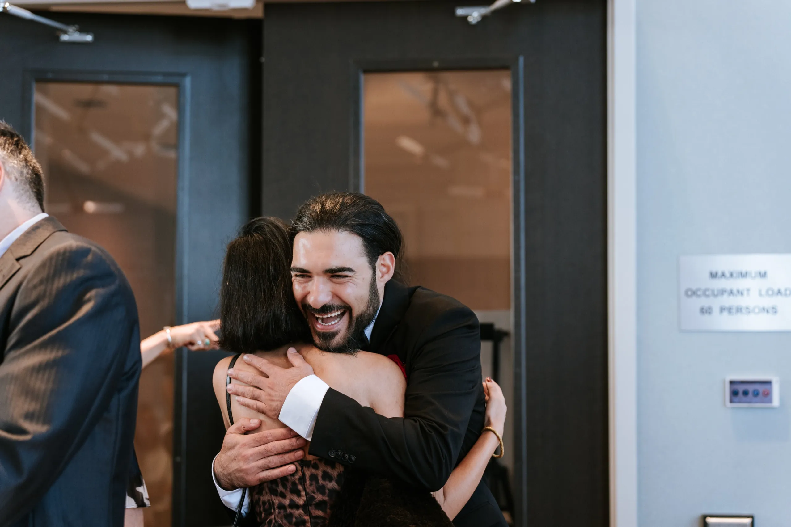 Moe laughing mid hug with an arriving guest at the party room doors in Maple, by AD Photography