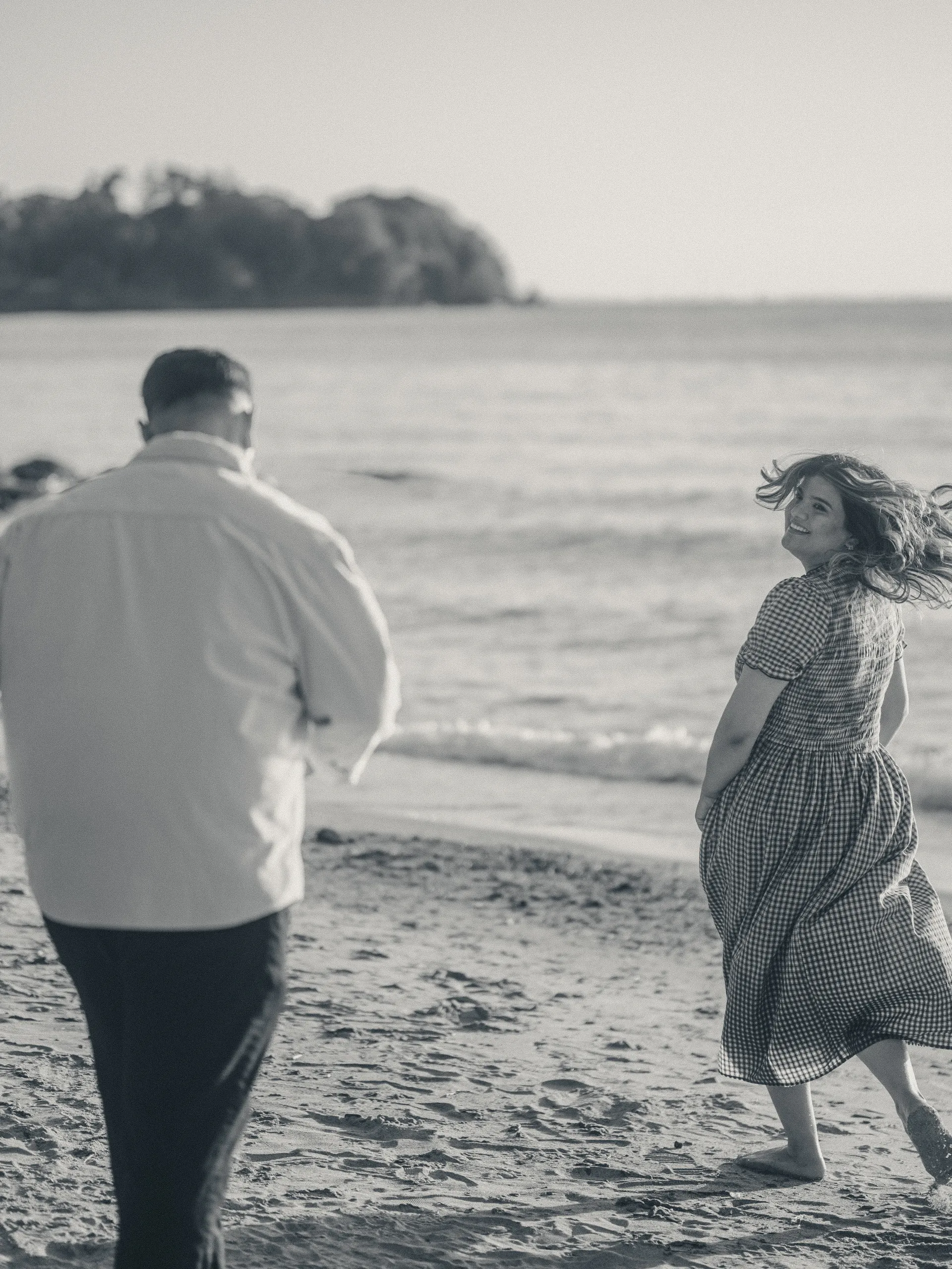 Black and white frame of Parneet running barefoot toward the water, hair flying, looking back at Maninder, by AD Photography