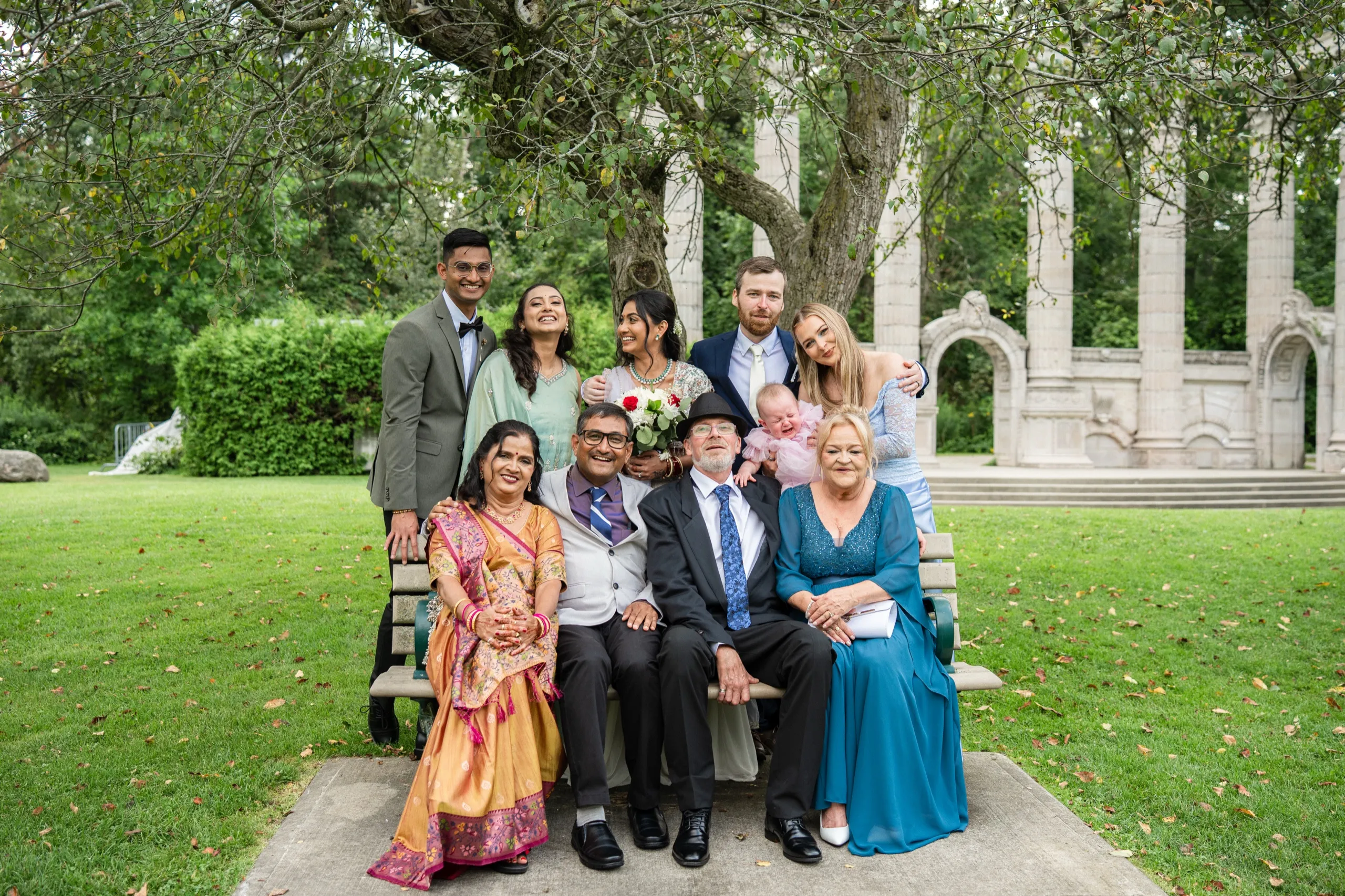 The extended family gathers on a bench beneath an old tree near the ceremony site., by AD Photography