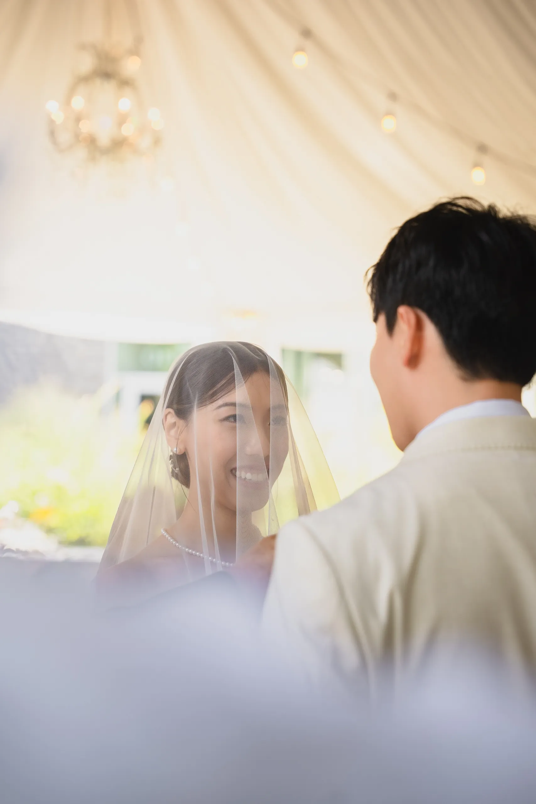 Josie smiling behind her veil in soft focus past Nelson's shoulder during the vows, by AD Photography
