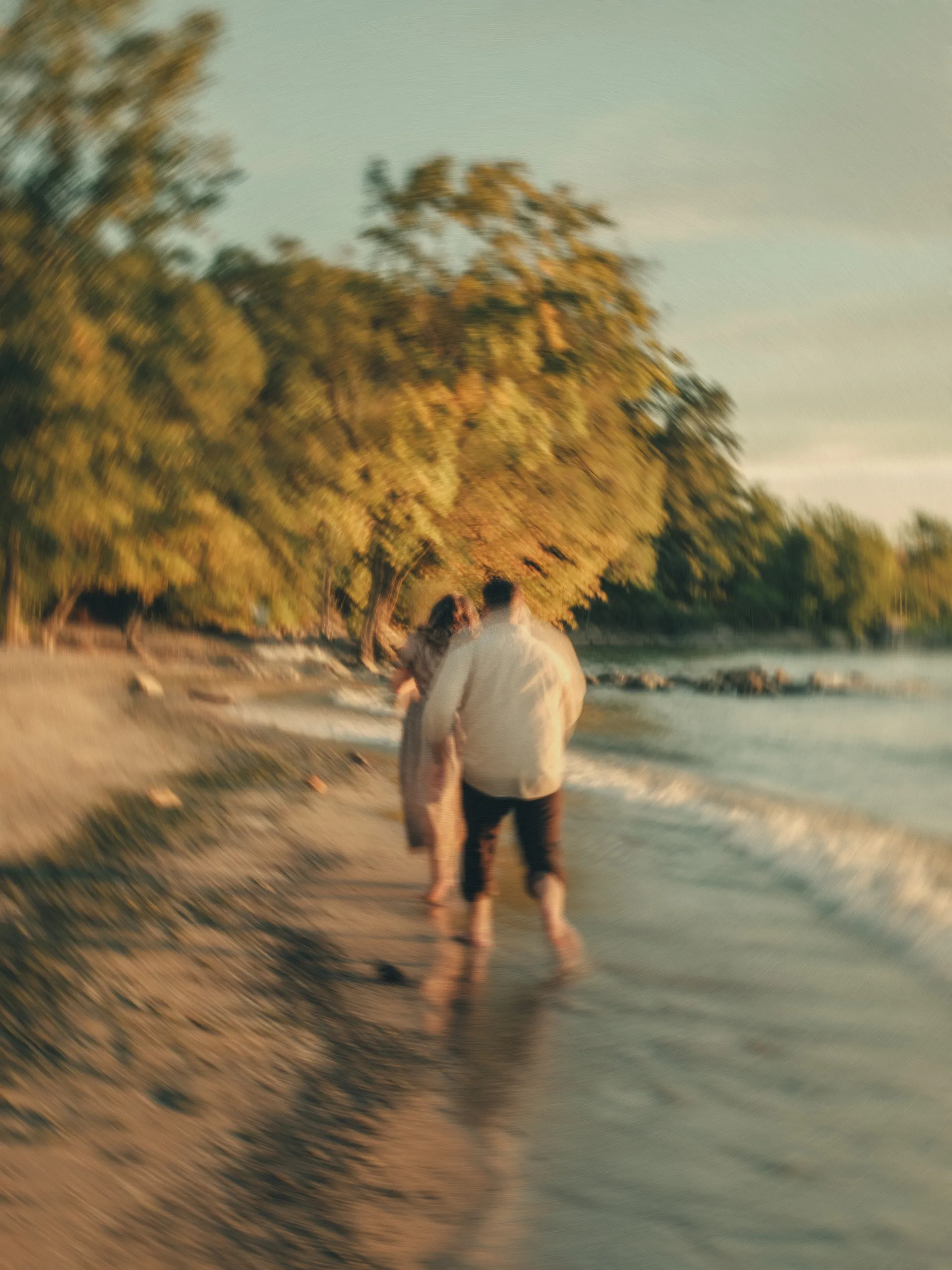 Maninder and Parneet walk away along the water's edge in soft blur, the shoreline trees lit gold, by AD Photography
