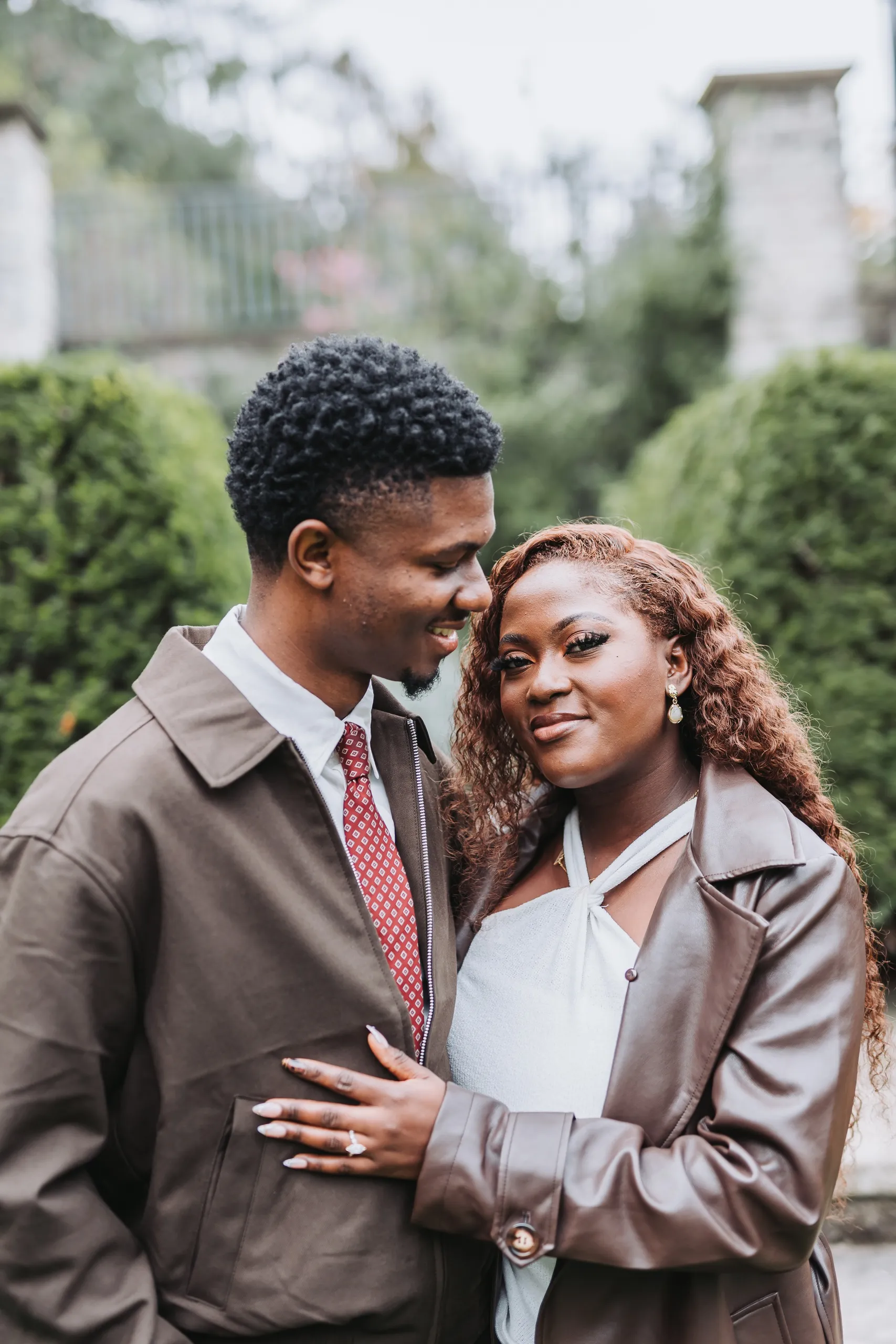 Aimee's ring hand on Richie's brown jacket as he smiles down at her between the clipped hedges, by AD Photography