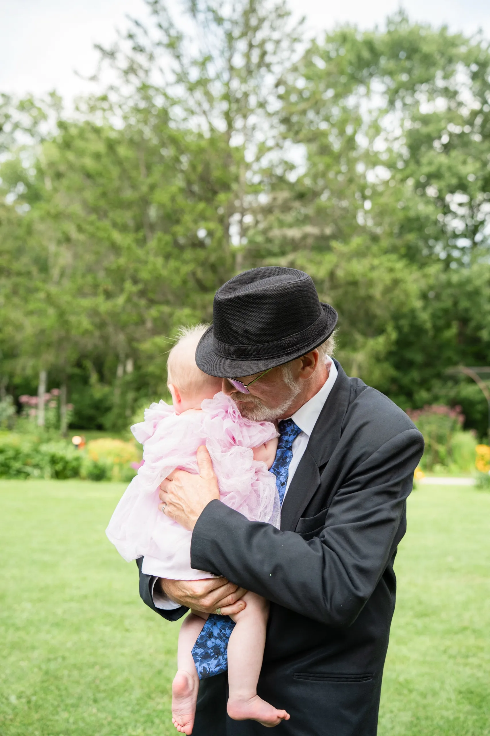 Derek's father cradles the flower girl close, his hat brim shading her tiny face., by AD Photography