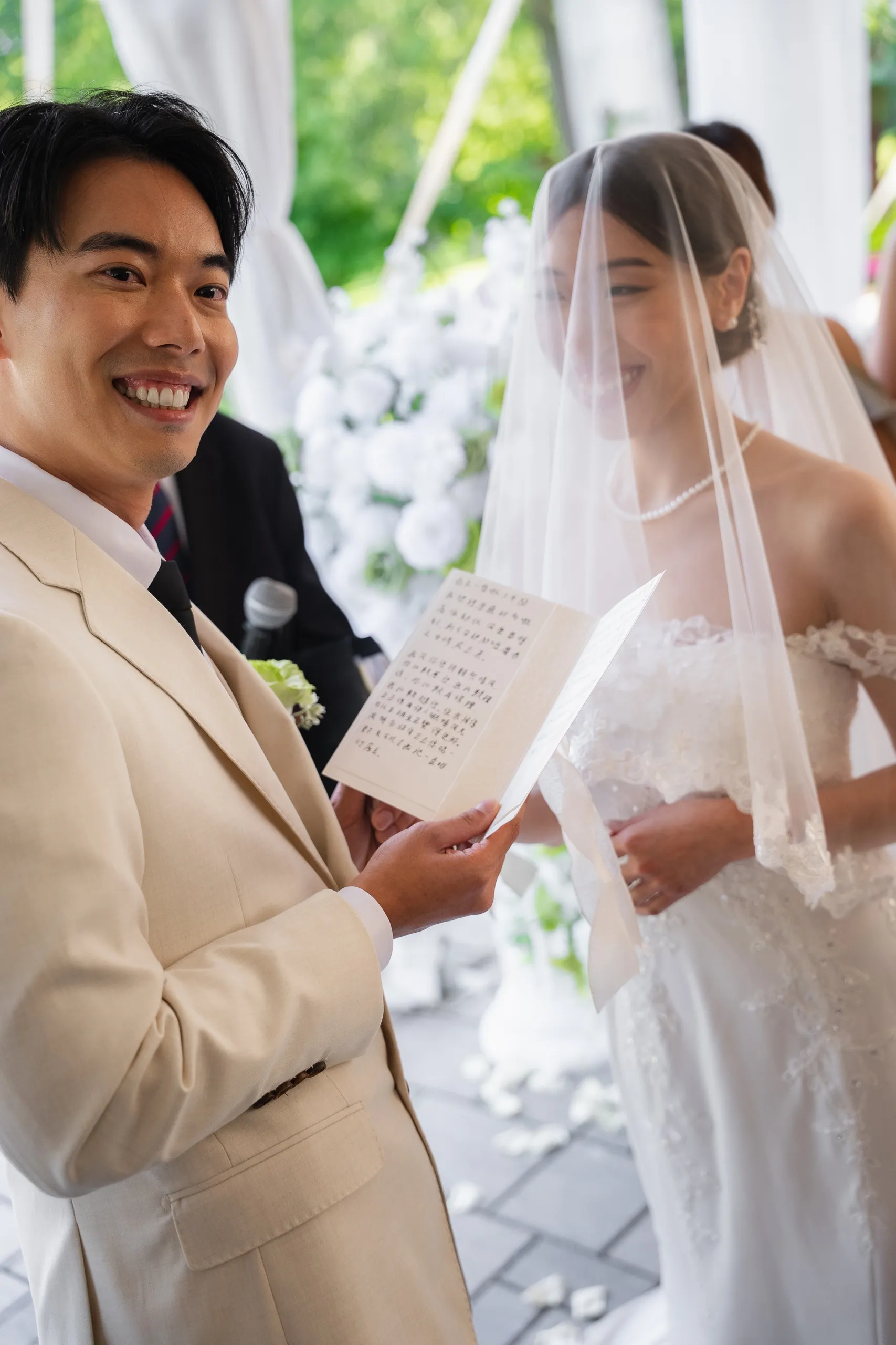 Nelson laughing mid vow with his card in hand while Josie beams behind her veil, by AD Photography