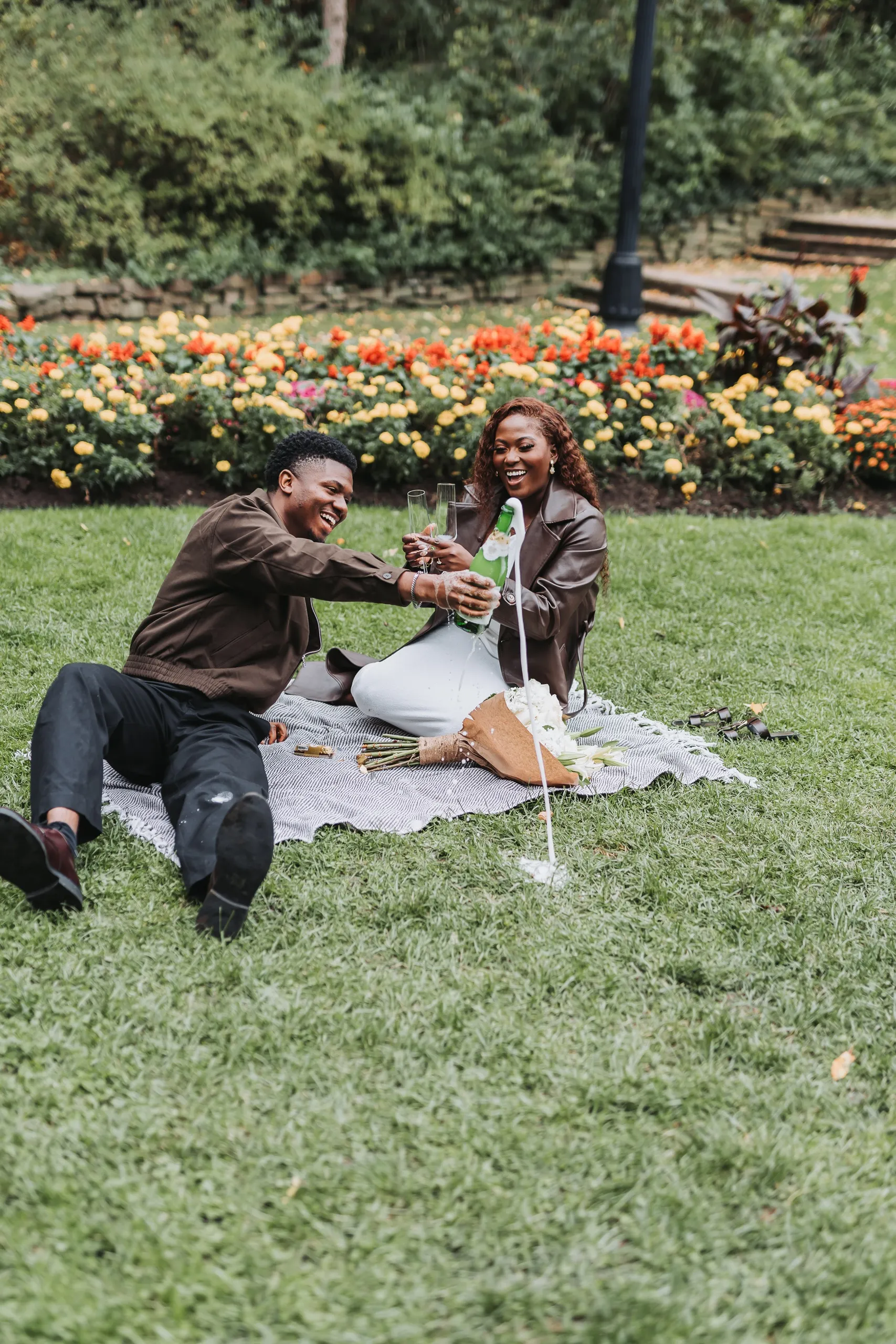 Richie pouring champagne on a picnic blanket as Aimee laughs with two glasses, by AD Photography
