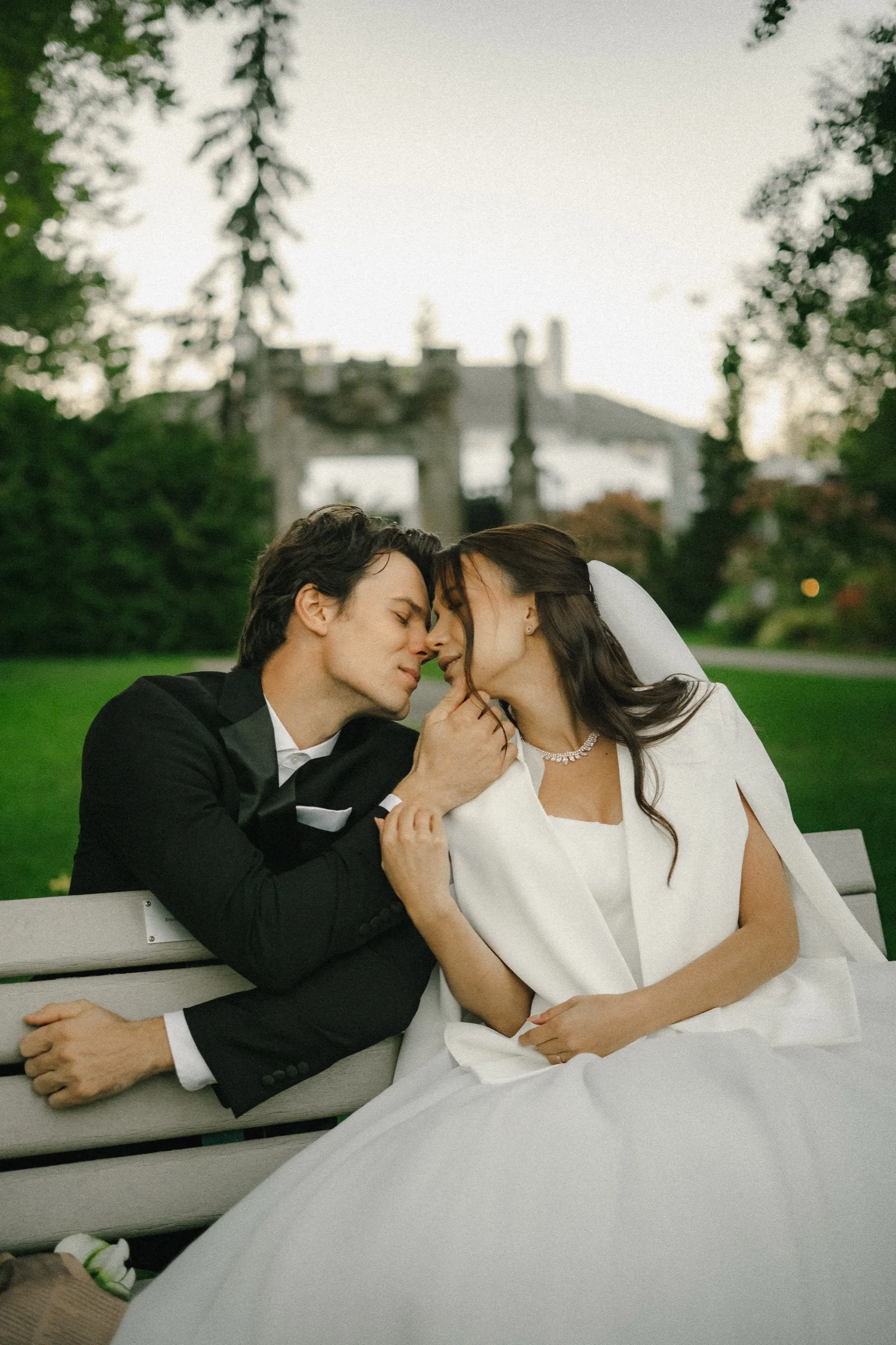 Bride in a white cape and groom nose to nose with their eyes closed on a white park bench, by AD Photography