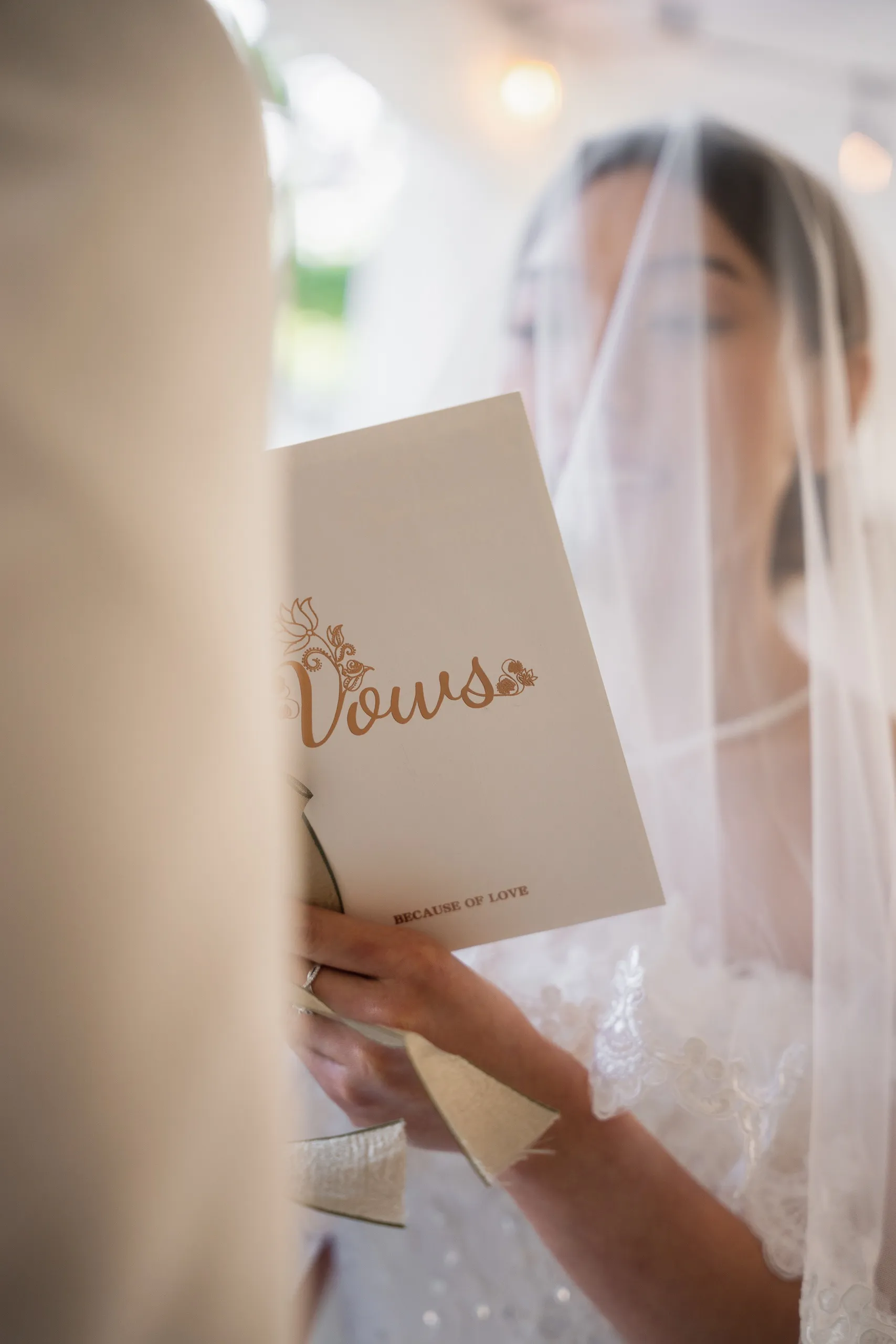 Josie's veiled hands holding the gold lettered vow booklet, framed past Nelson's shoulder, by AD Photography