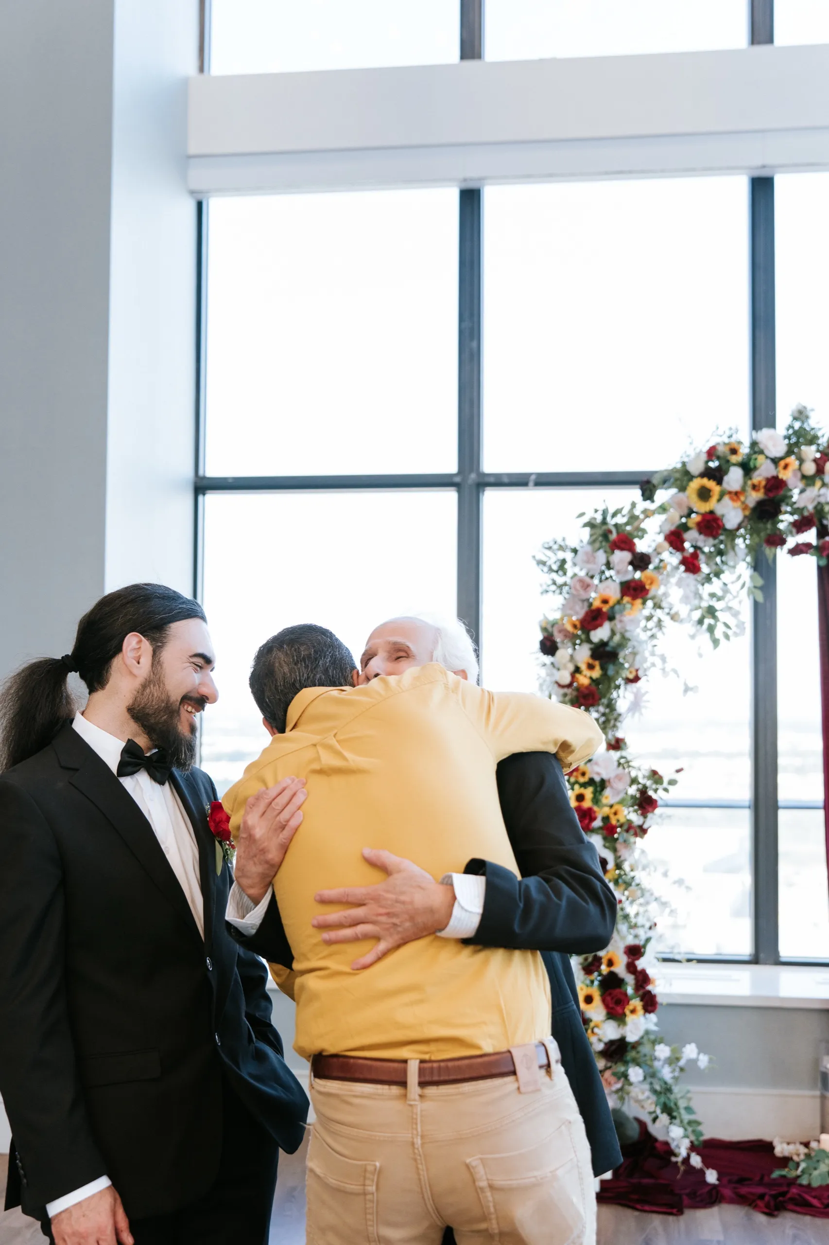 A guest in yellow embraces a family elder in front of the sunflower arch while Moe smiles alongside, by AD Photography