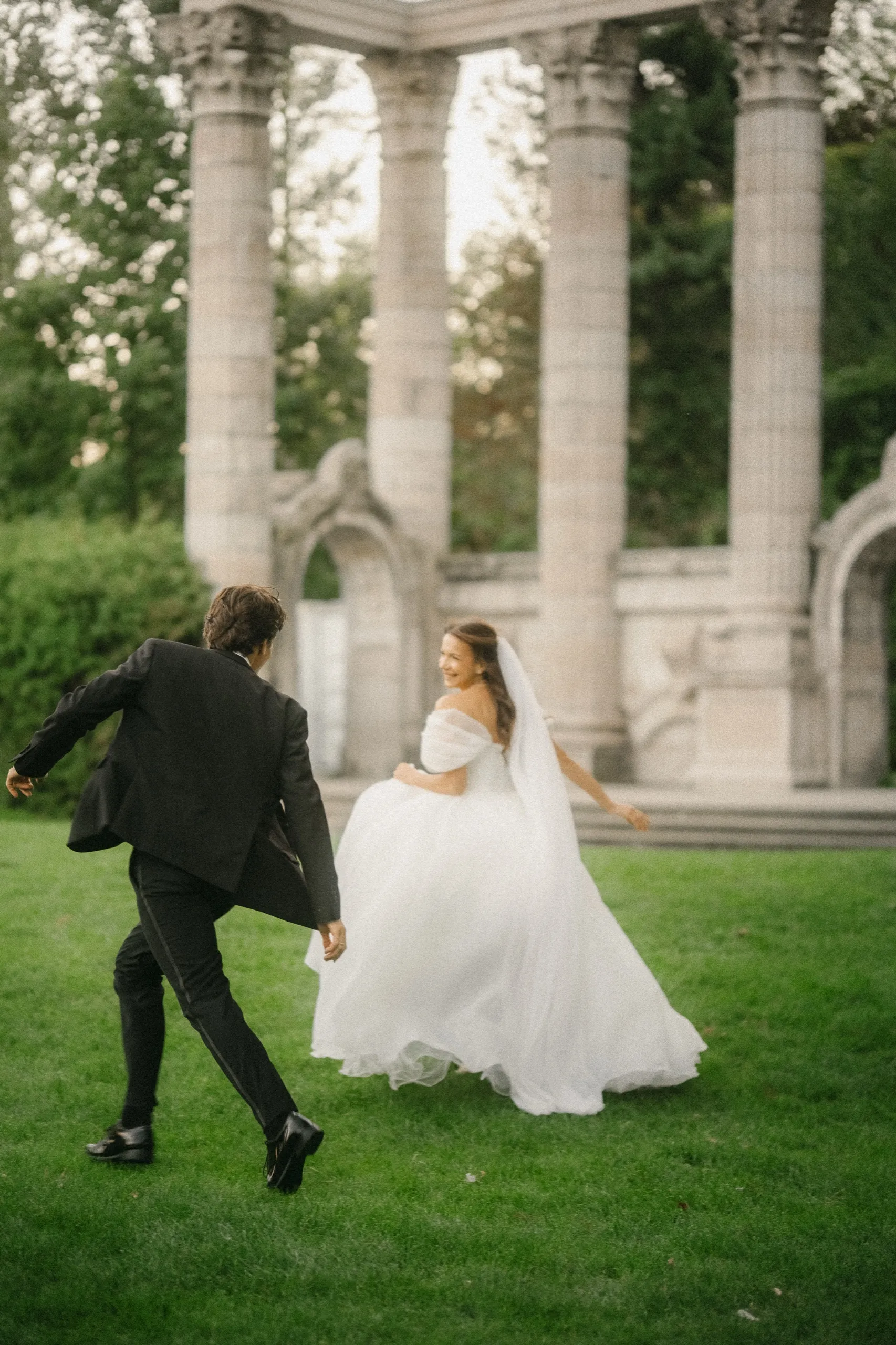 Groom chasing the bride across the lawn as she looks back laughing below the colonnade, by AD Photography