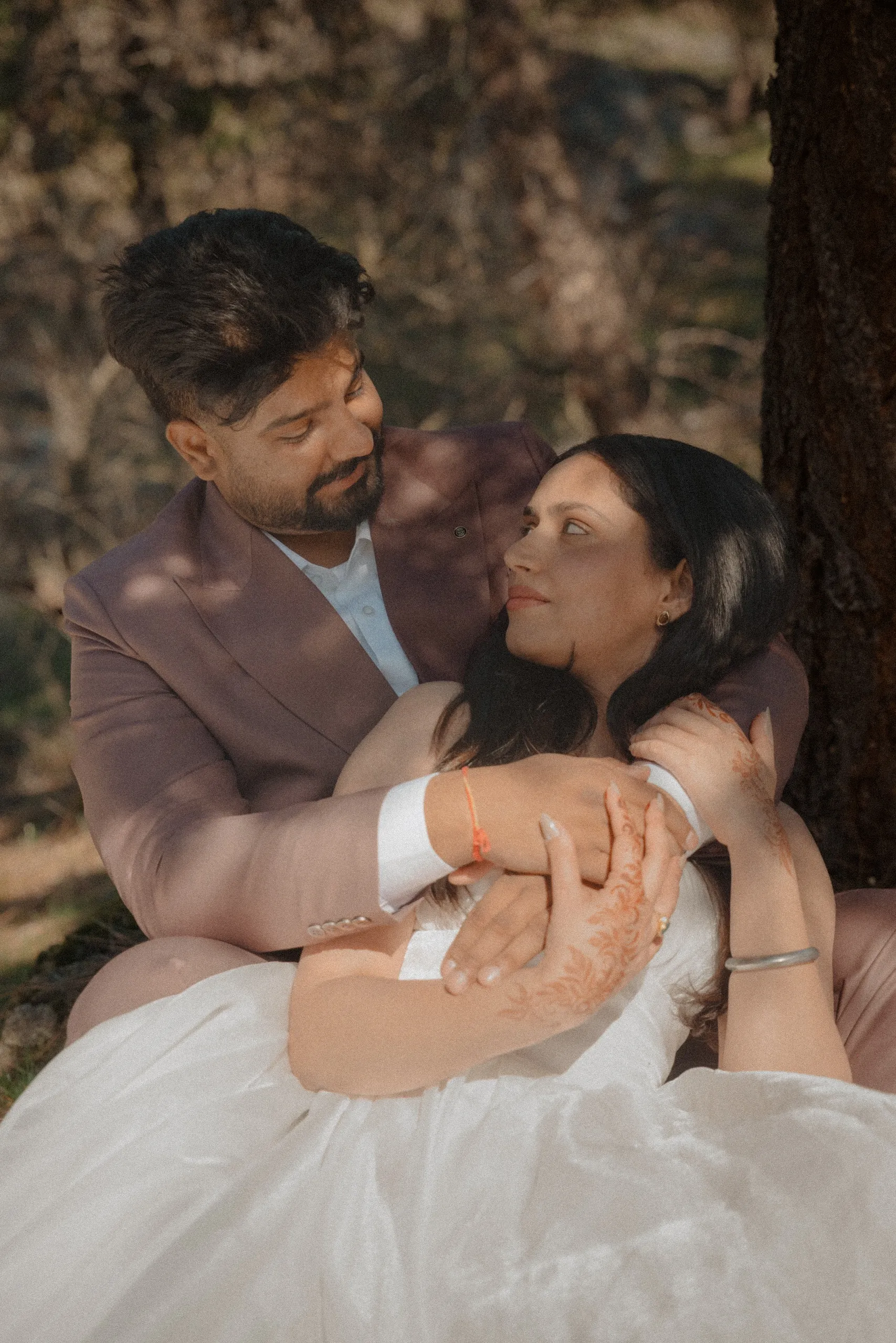 The couple sitting under a pine, his arms wrapped over her henna covered hands, by AD Photography