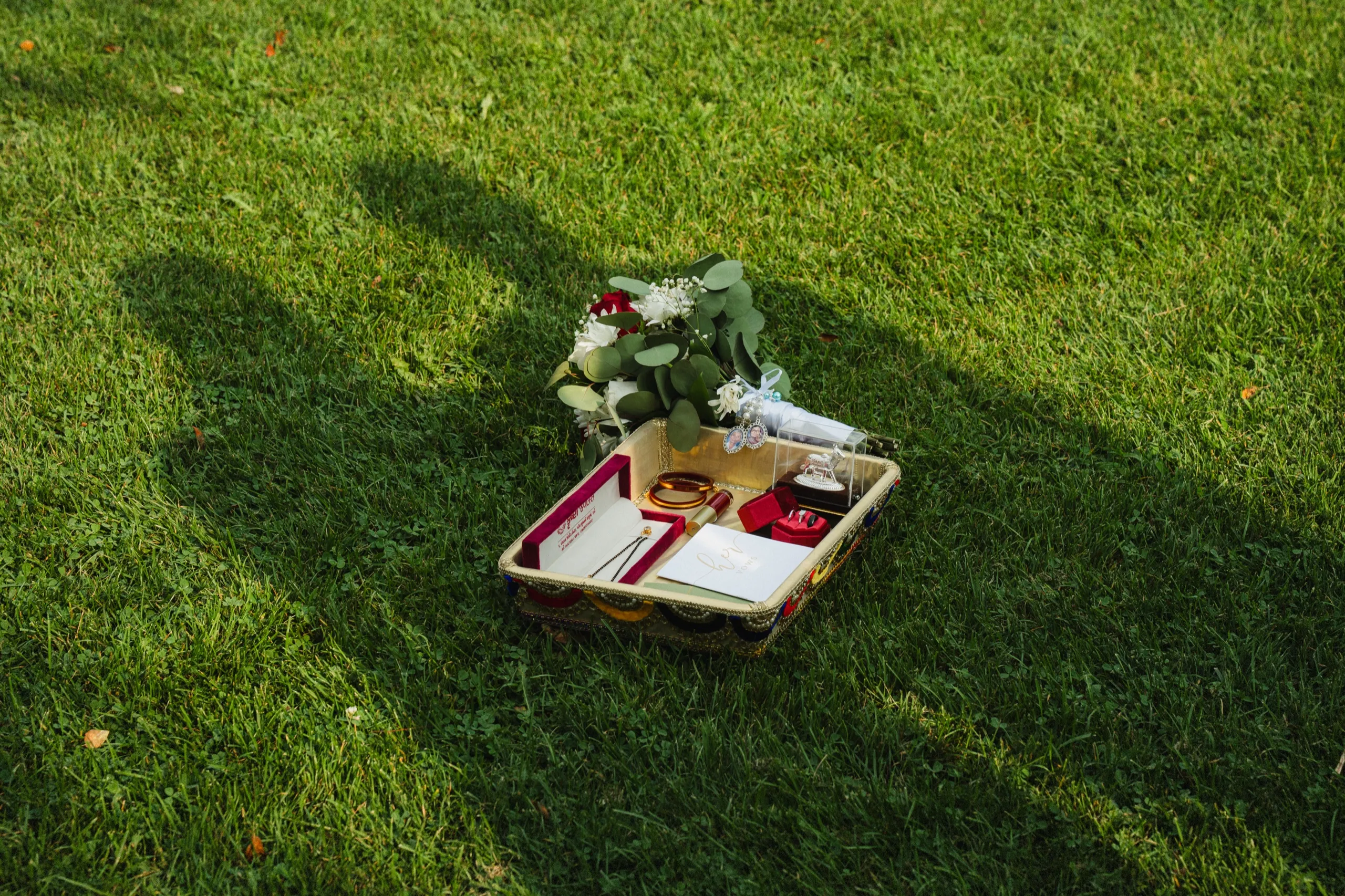 A gold tray holds bangles, lipstick, jewelry boxes, and the bouquet laid on the grass., by AD Photography