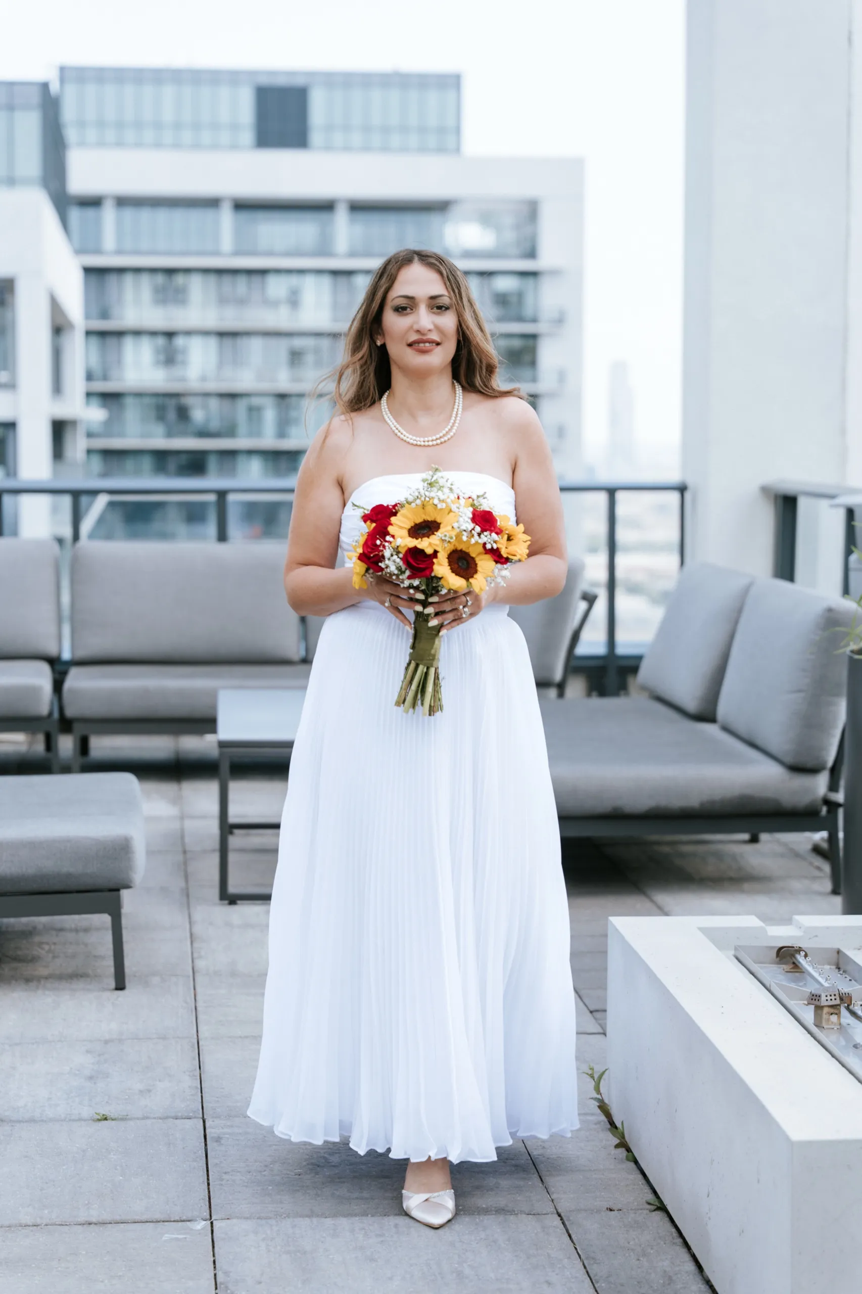 Zoha on the terrace in her white pleated gown with a sunflower bouquet, Maple condo towers behind, by AD Photography