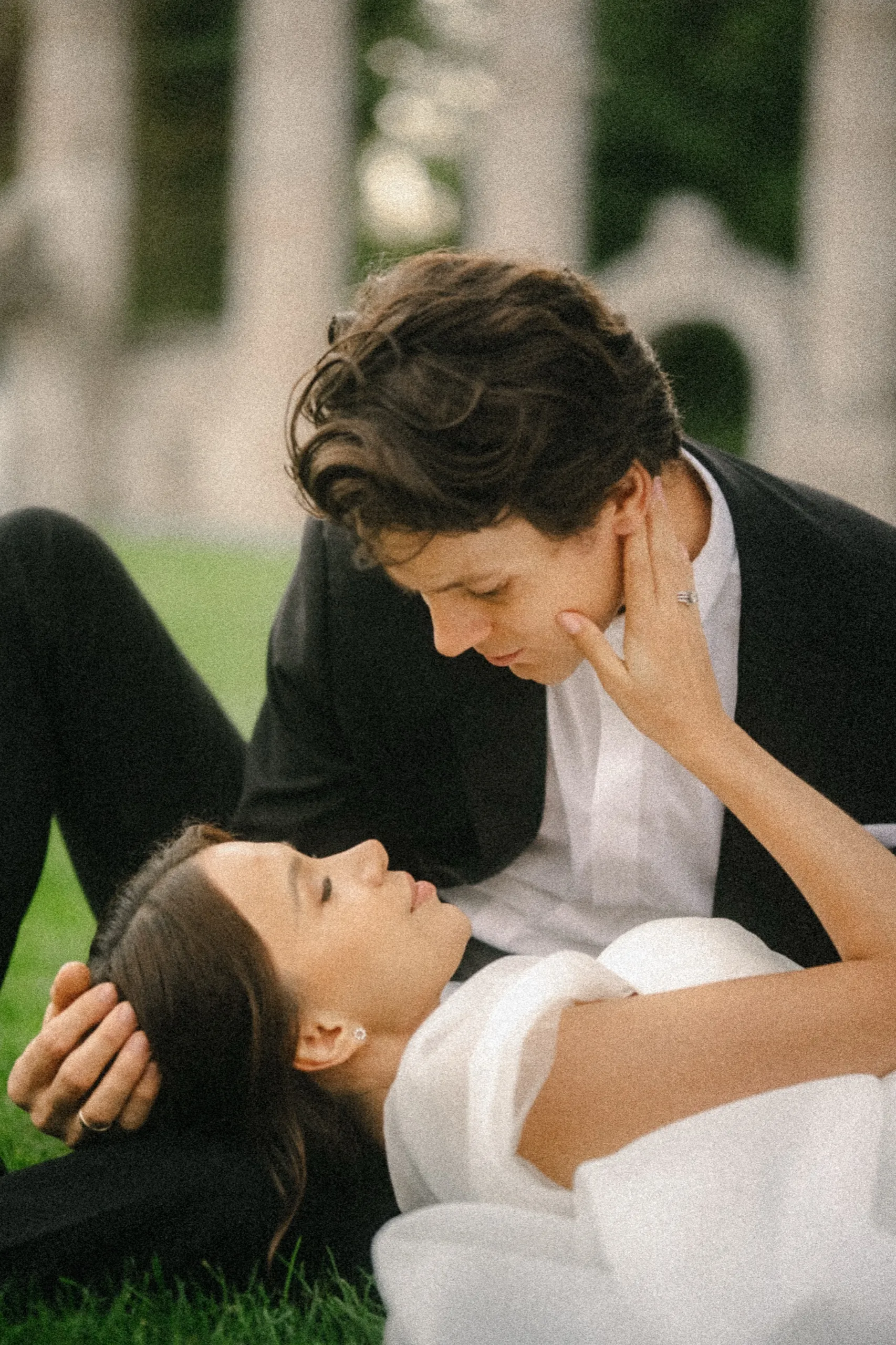 Bride lying in the grass touching the groom's jaw as he leans down for an almost kiss, by AD Photography