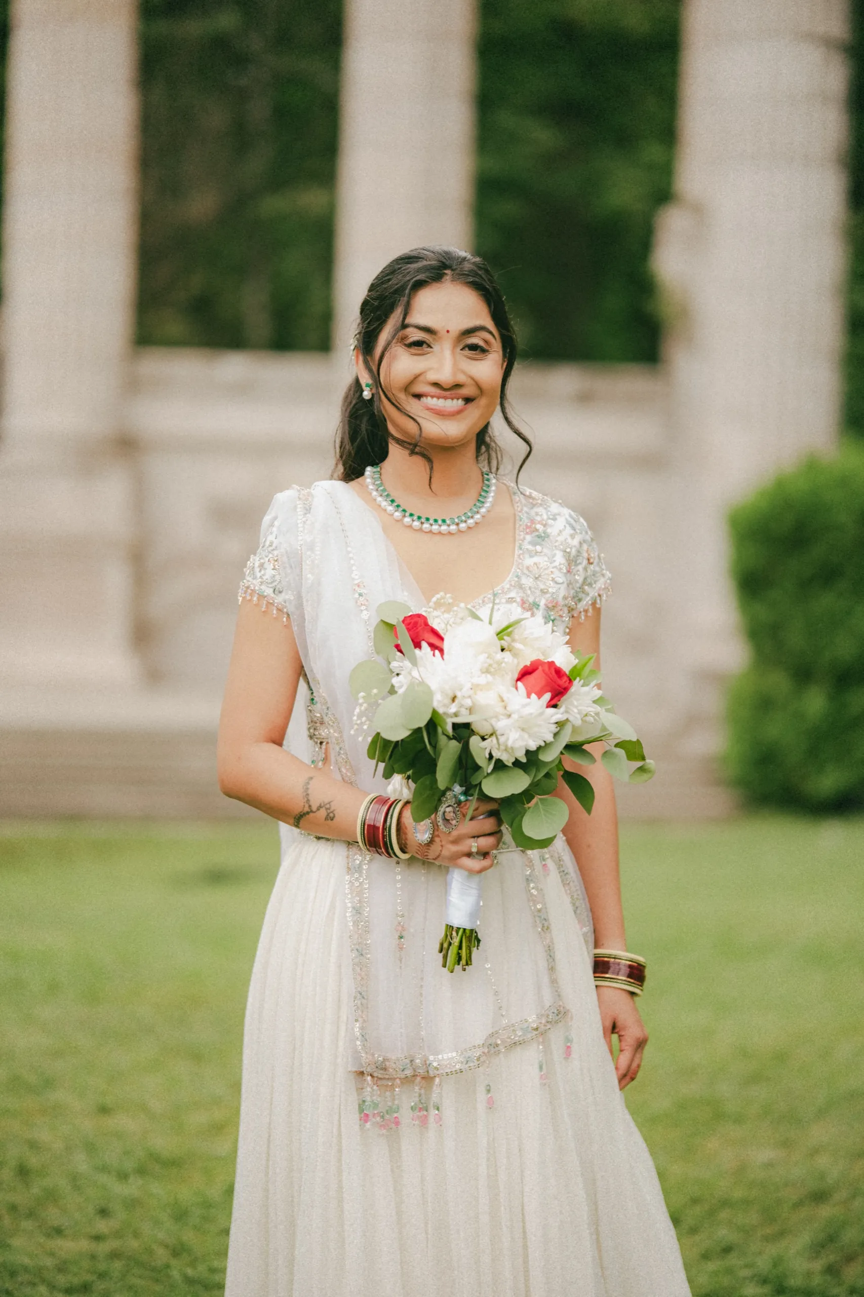 Nidhi smiles holding her bouquet of red and white blooms in front of the columns., by AD Photography