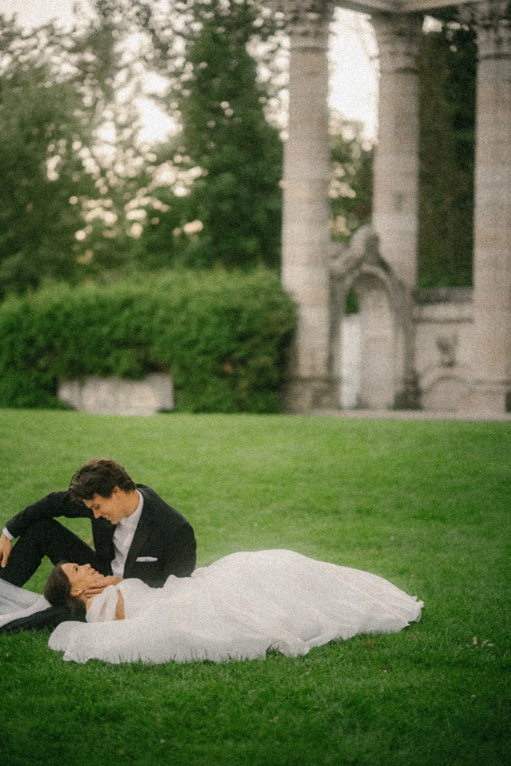 Bride's gown pooled across the lawn as the groom leans over her, columns and carved arch behind, at Guild Park at sunset, by AD Photography