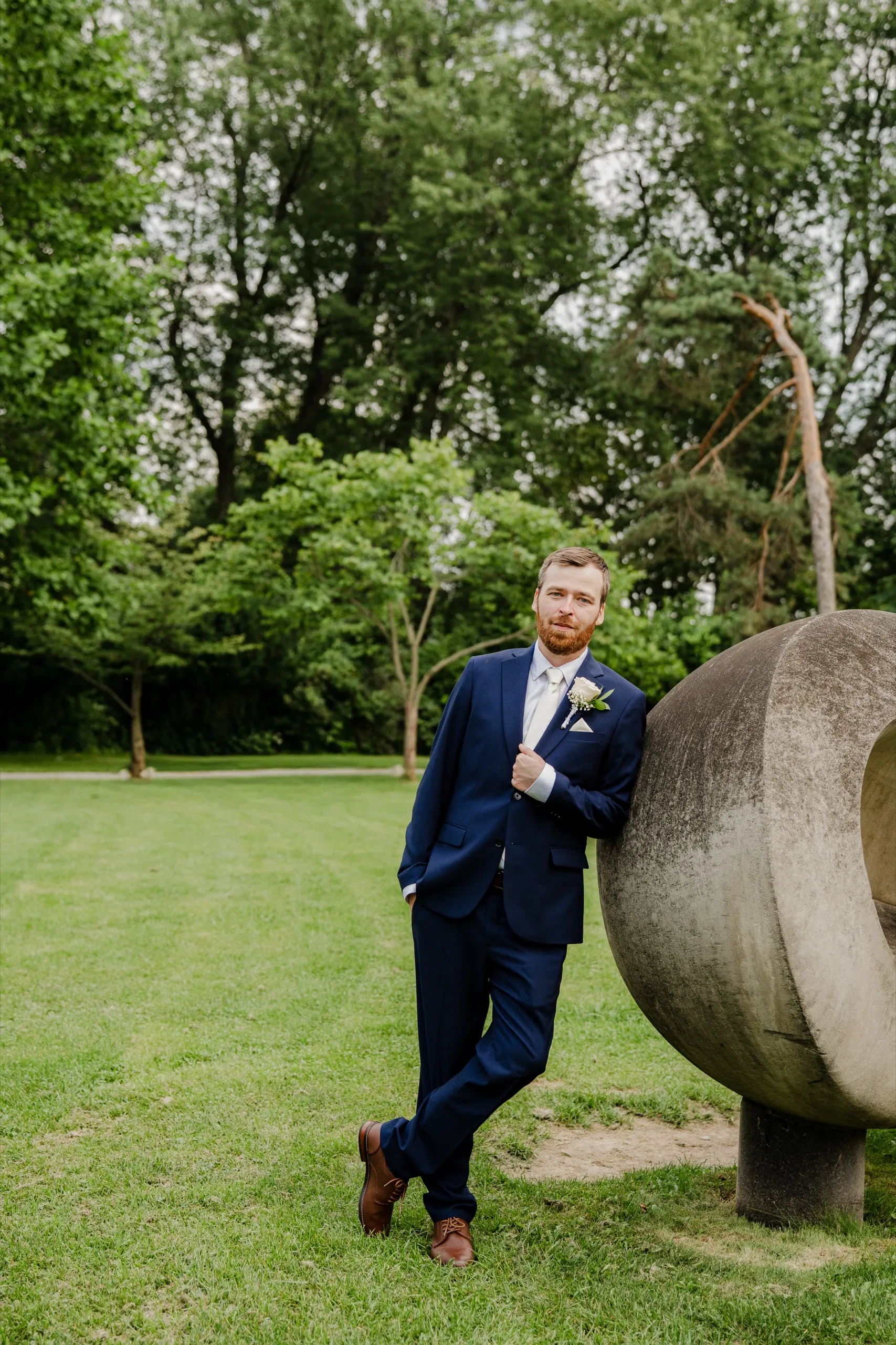 Derek leans against a large stone sculpture on the open lawn in his navy suit., by AD Photography