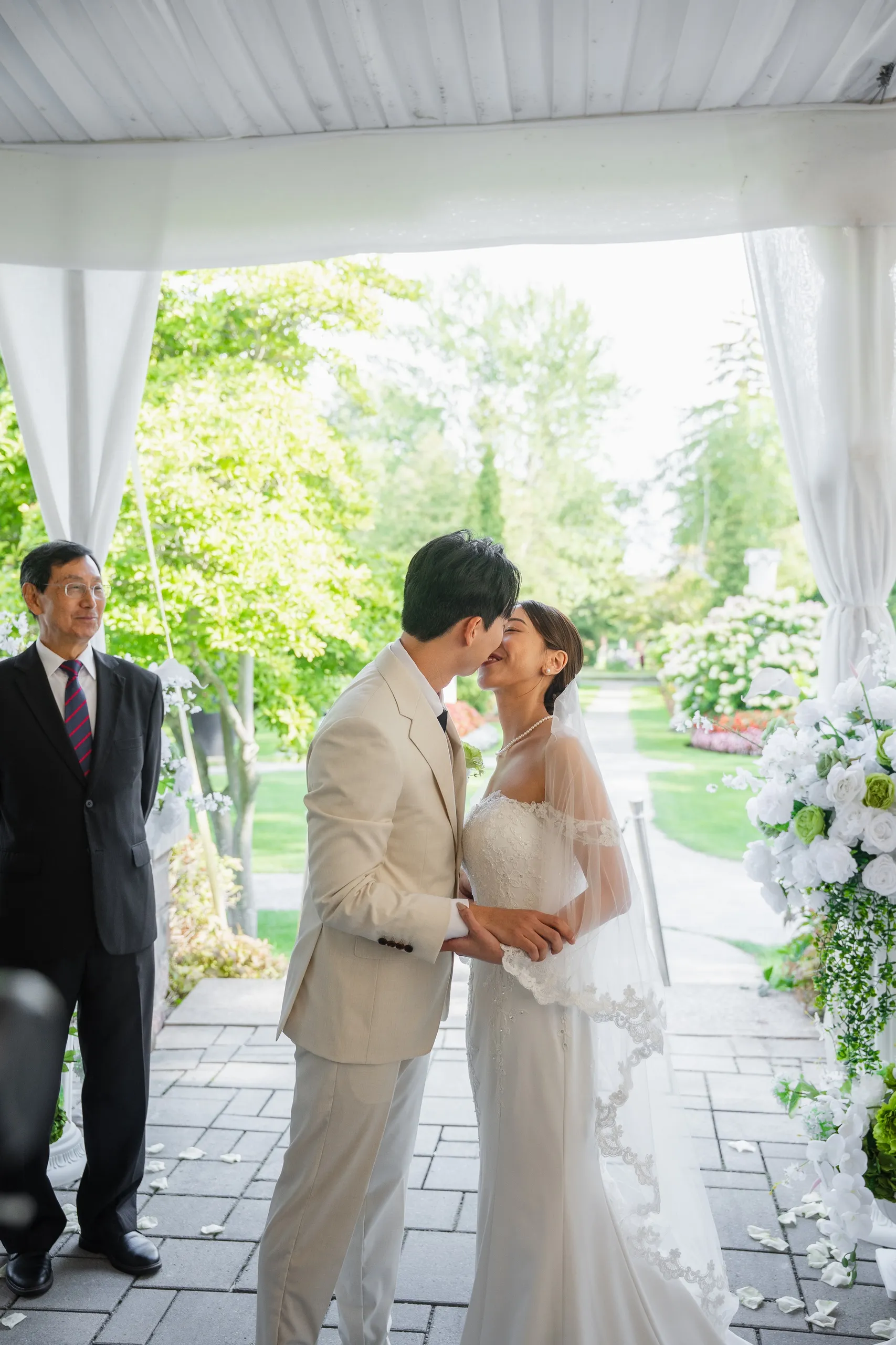 First kiss as the officiant looks on, garden and petal strewn stone behind them, by AD Photography