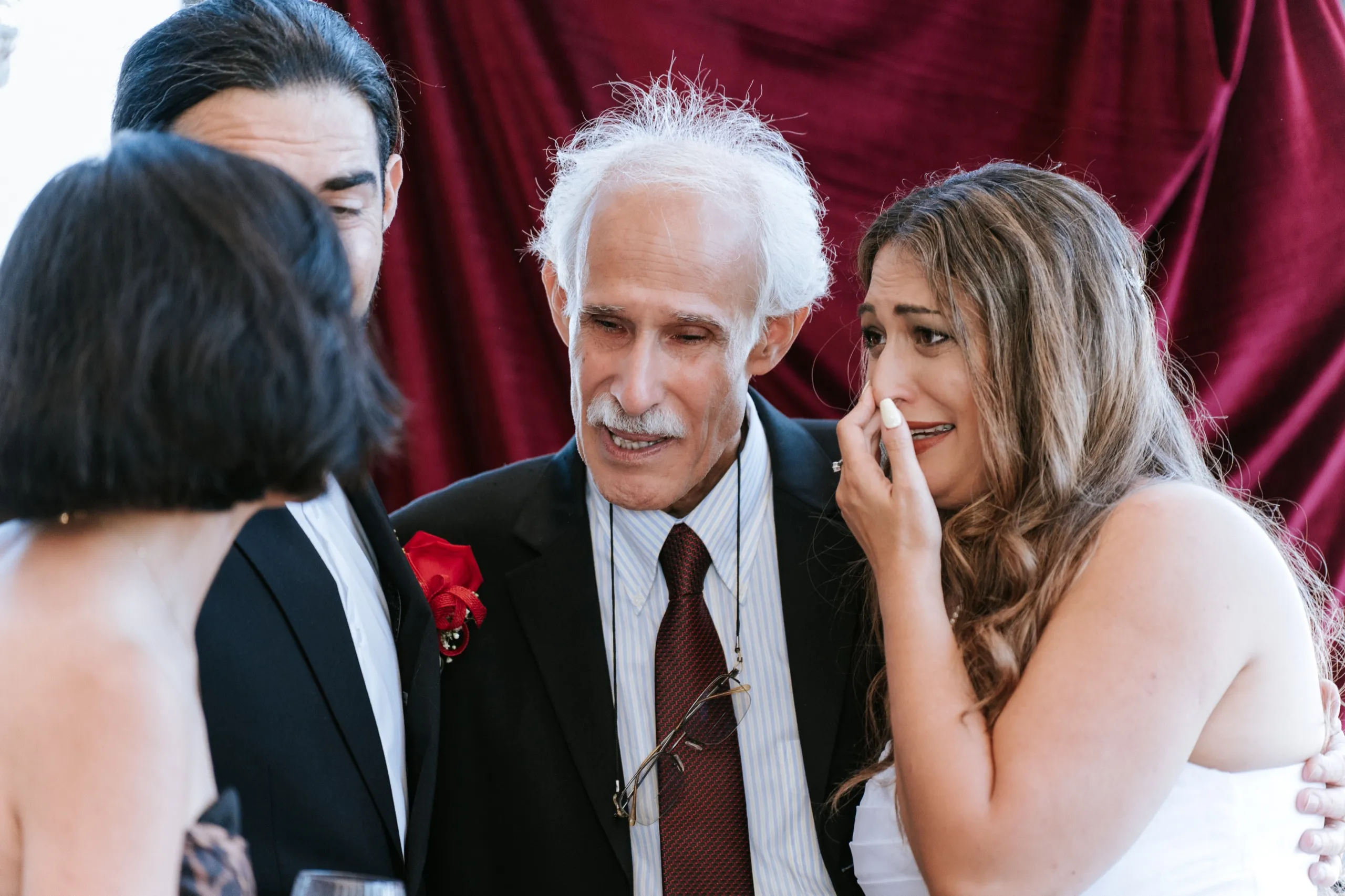 Zoha wipes her eyes beside a family elder in front of the burgundy drape, by AD Photography