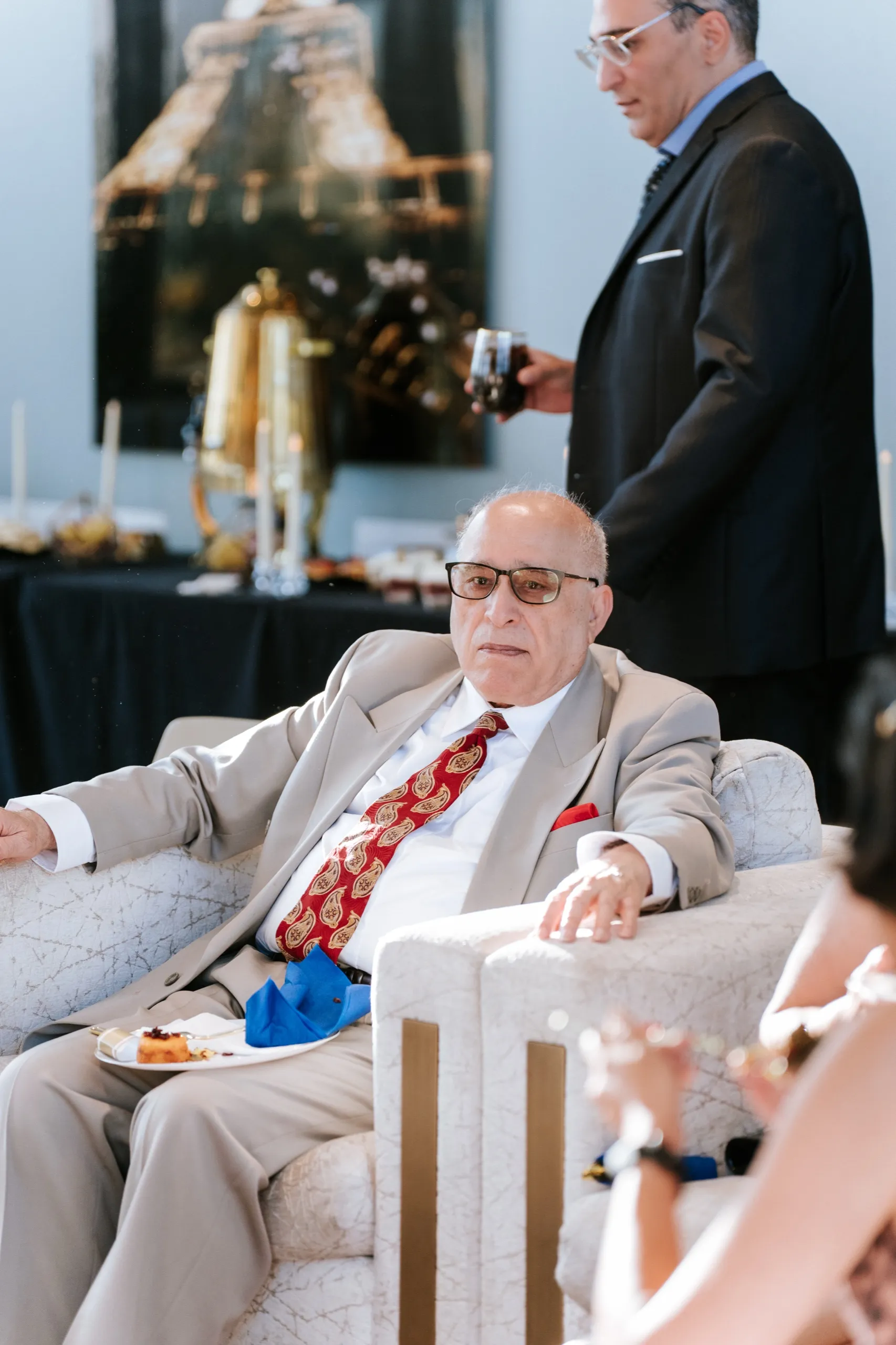 A guest in a tan suit sits back with a plate of dessert in the lounge chair, by AD Photography