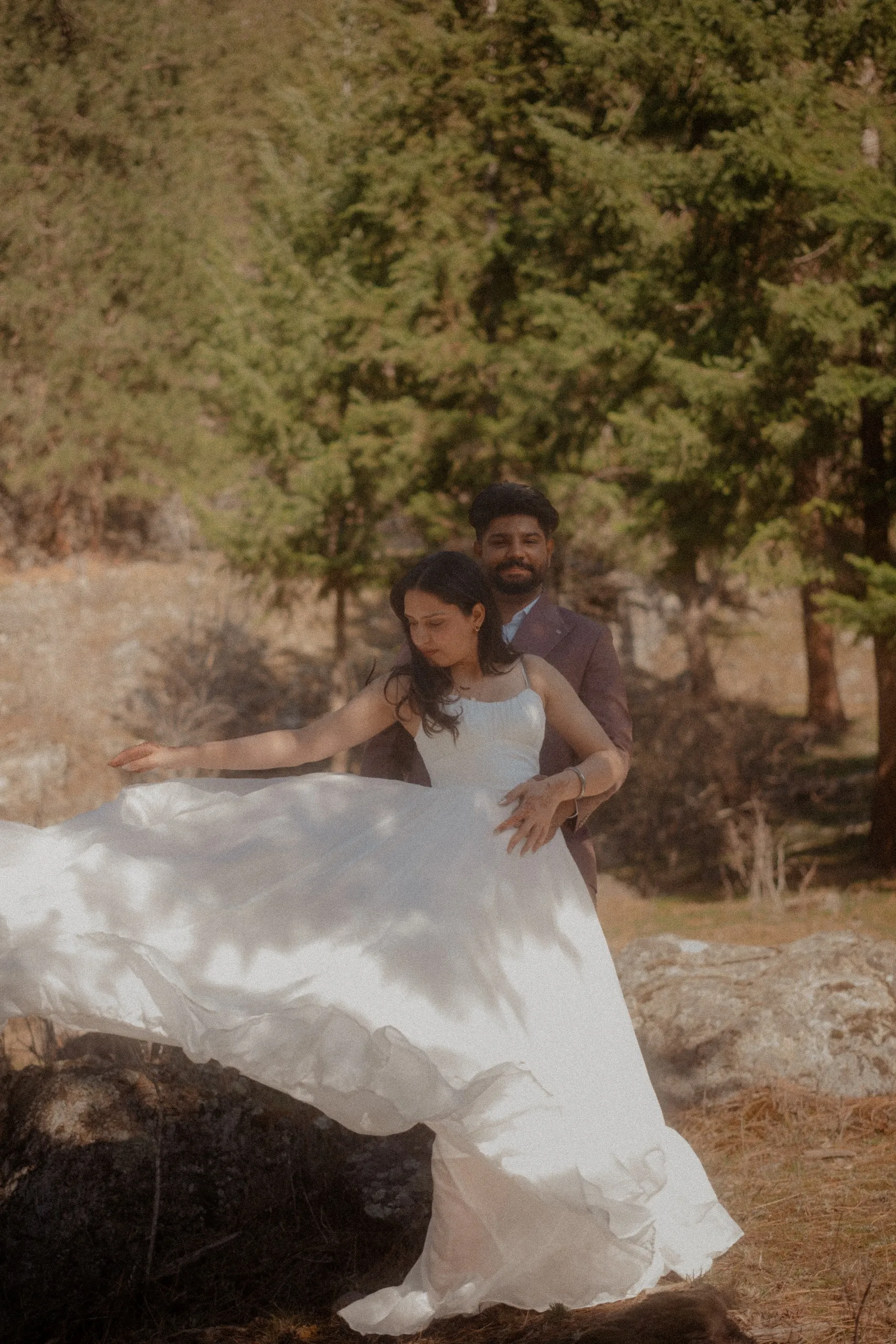 The bride dragging her gown across dry grass with Kamal watching from the pines, by AD Photography